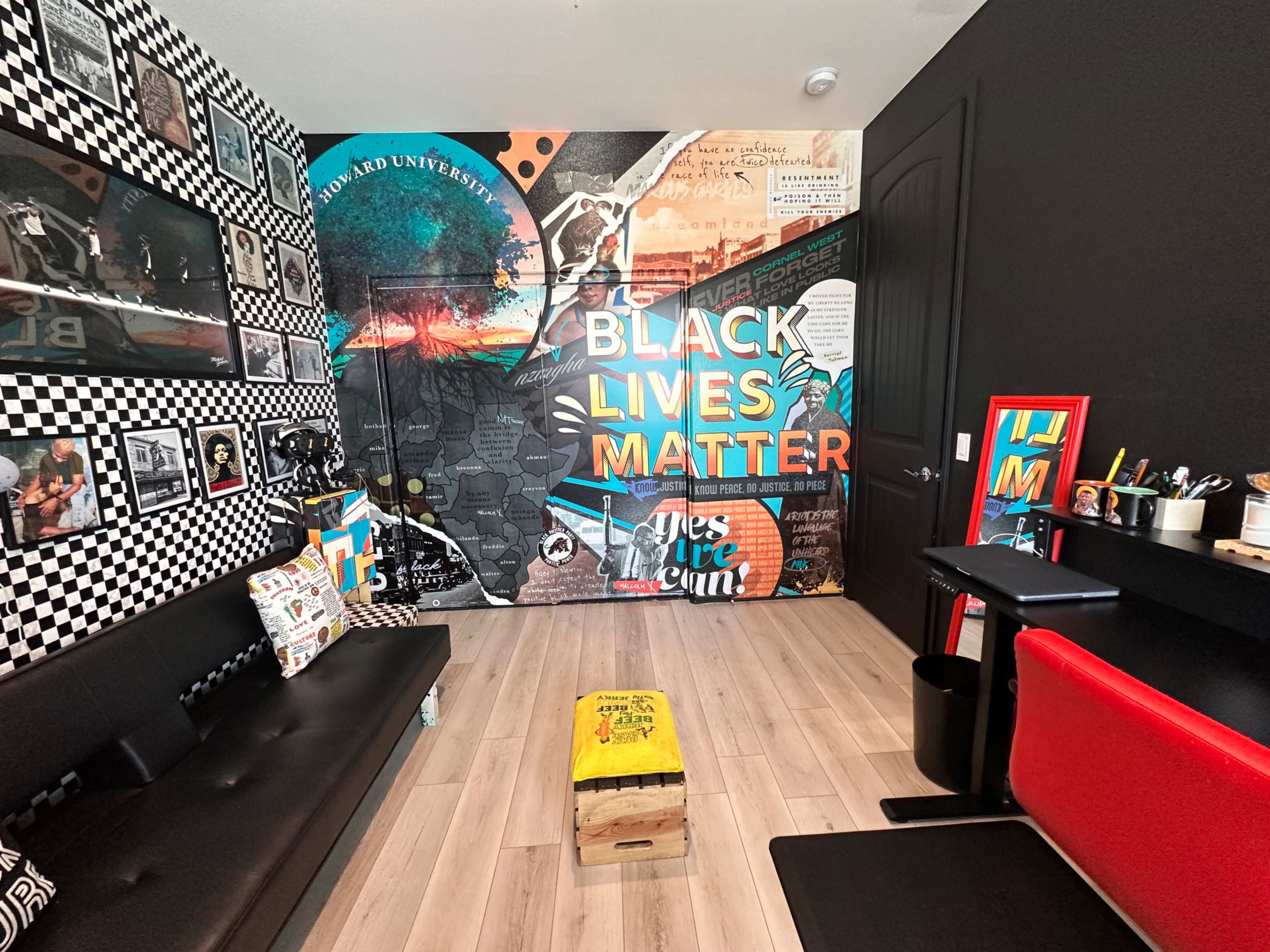 A room with a mural reading "BLACK LIVES MATTER" on one wall, accompanied by various framed artworks and a black sofa facing a small table and counter.