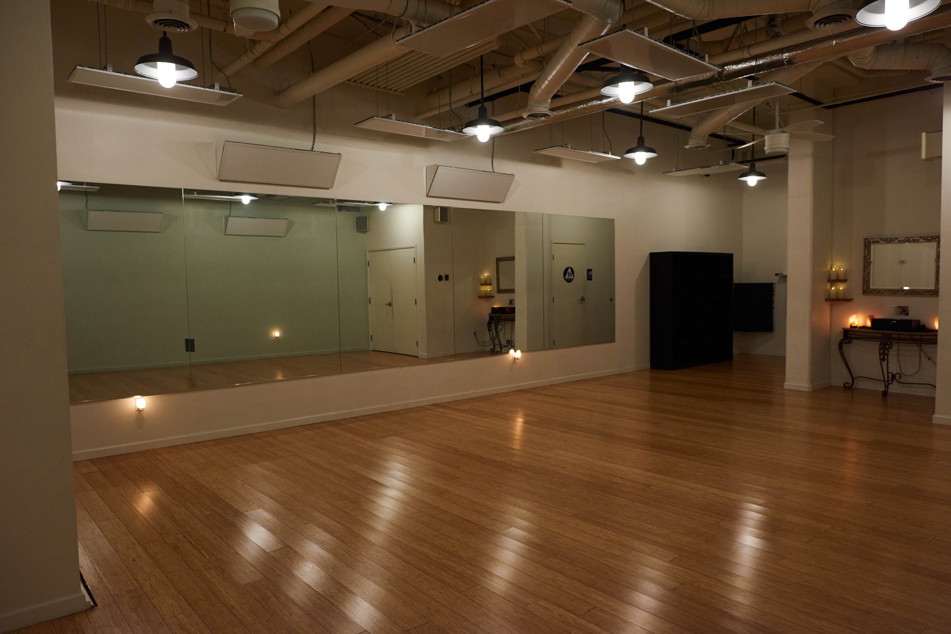 A spacious dance studio with a wooden floor, large mirrors on one wall, and minimal furnishings.