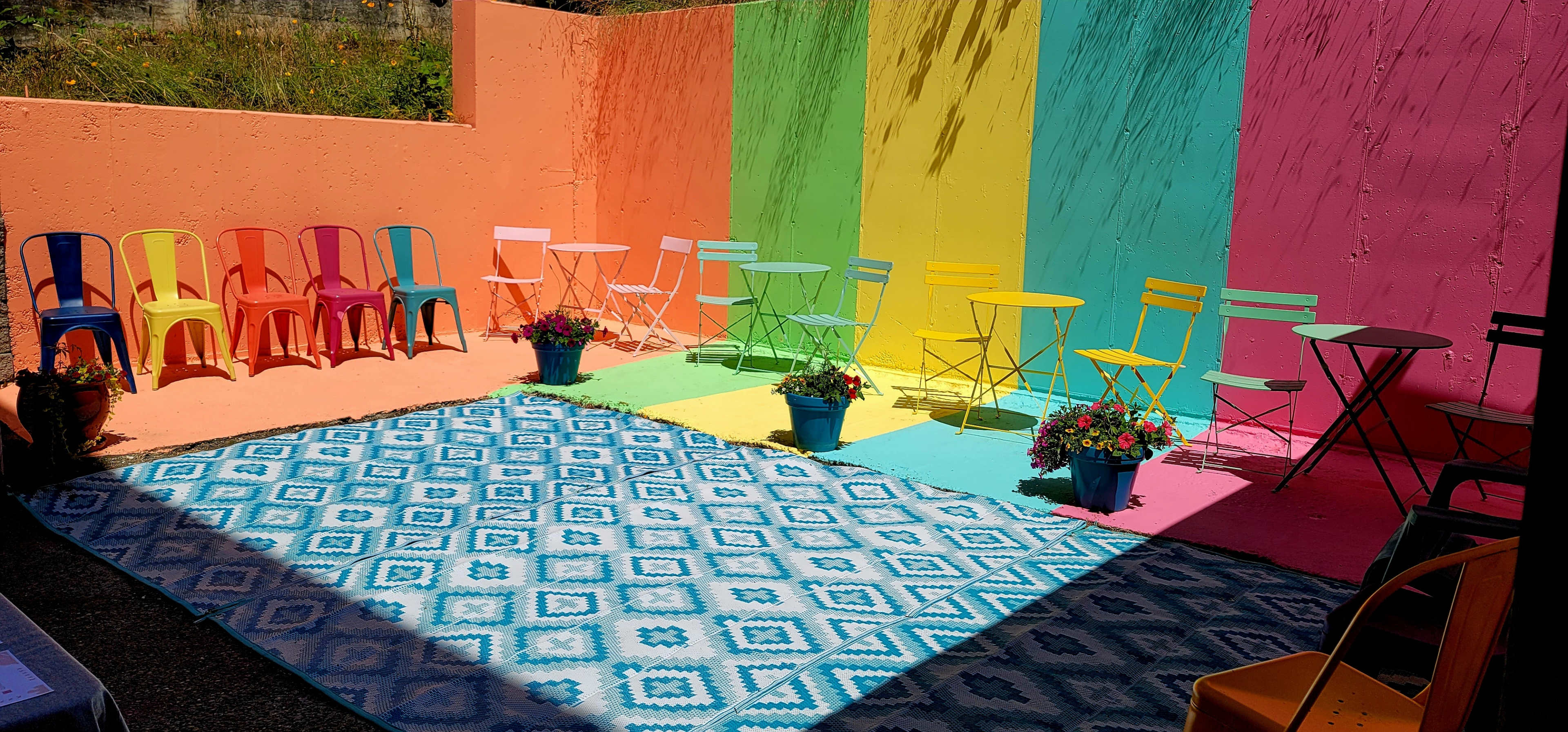 The scene features a brightly colored outdoor space with chairs and tables arranged around a patterned rug and vibrant walls.