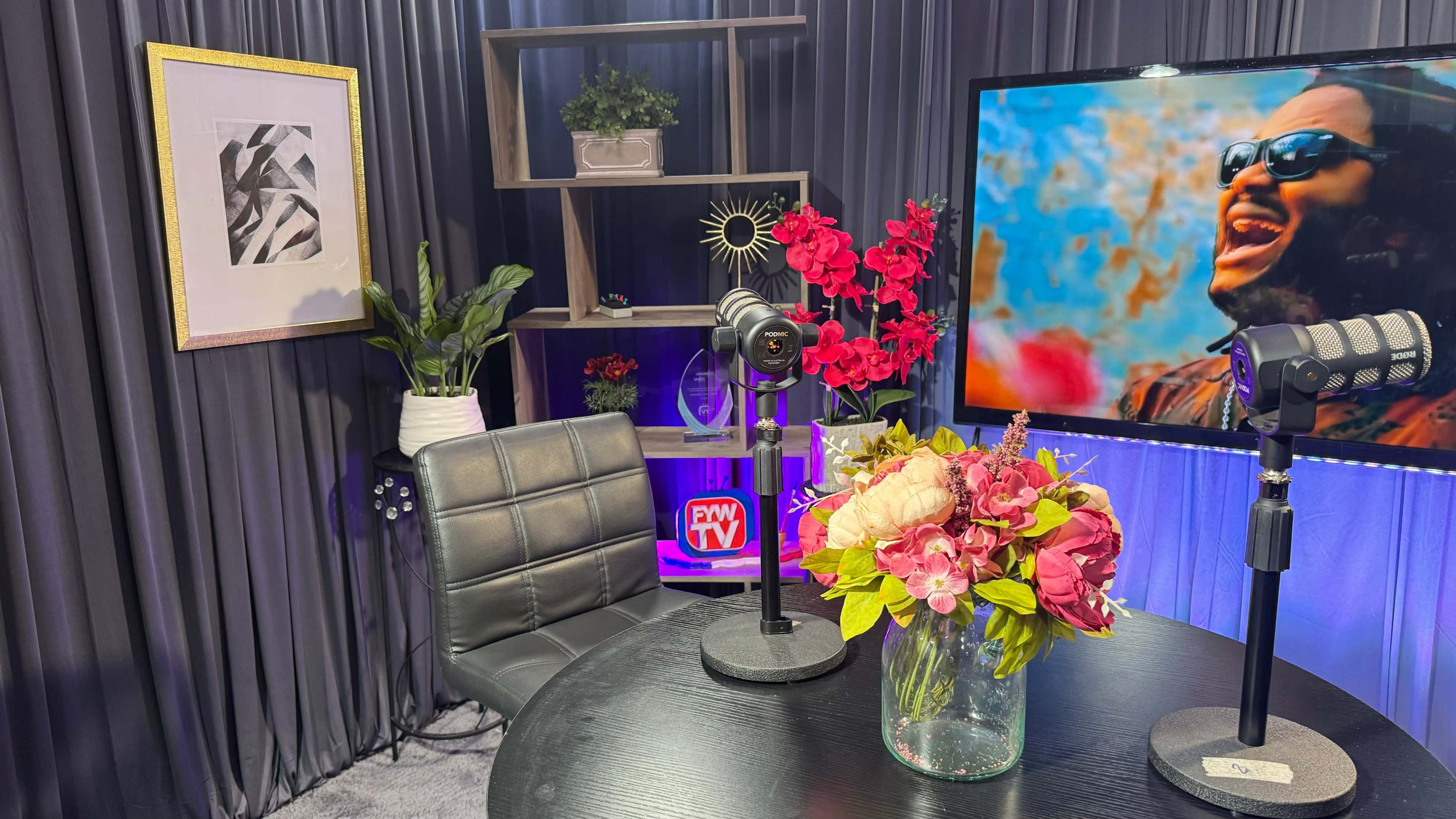 The image shows a modern studio setup with a round table, a microphone on each side, a floral arrangement in a vase, and a large screen displaying a colorful image in the background.