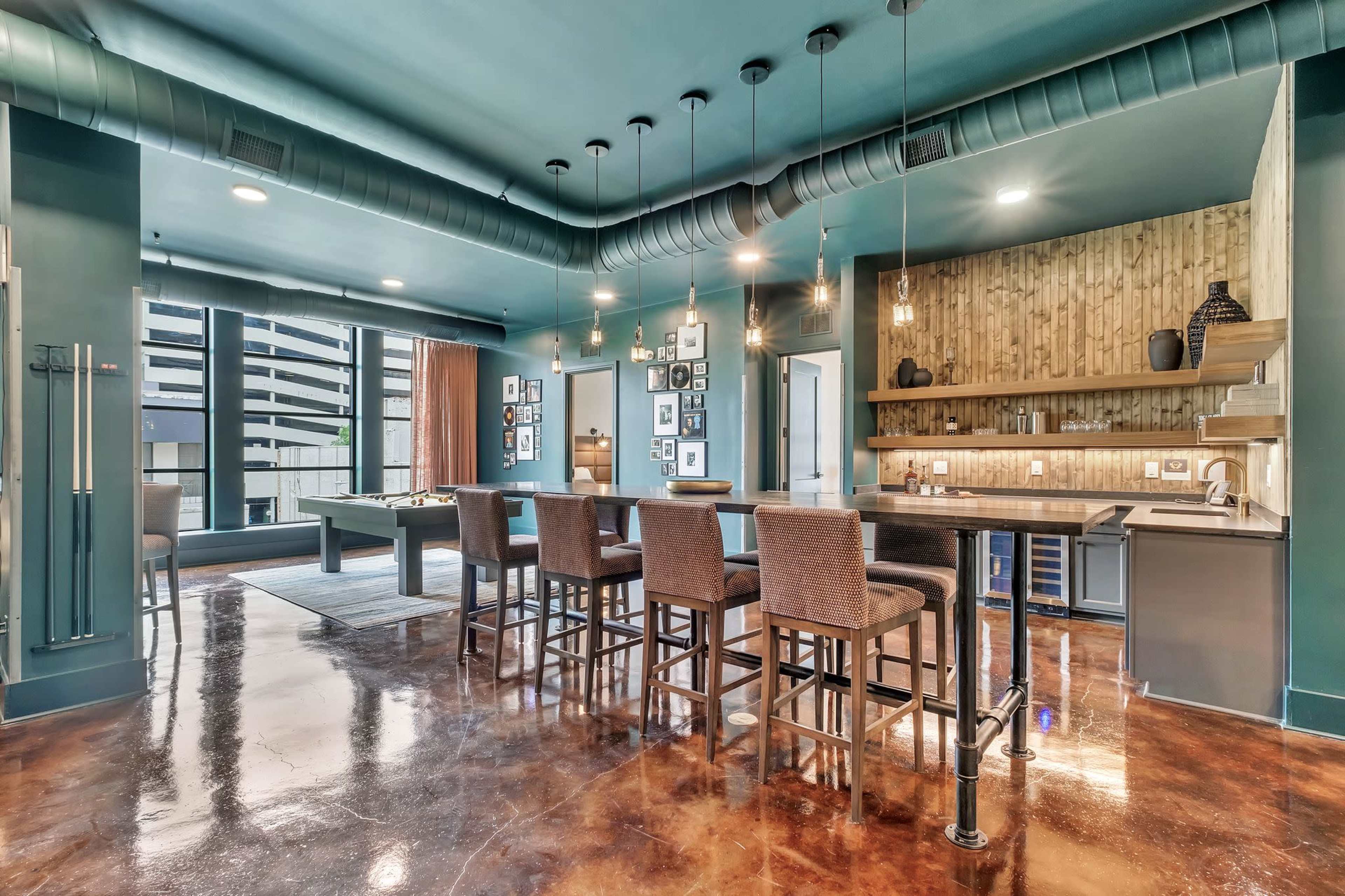 The image shows a modern lounge area featuring a bar with high chairs, polished concrete floors, and large windows.