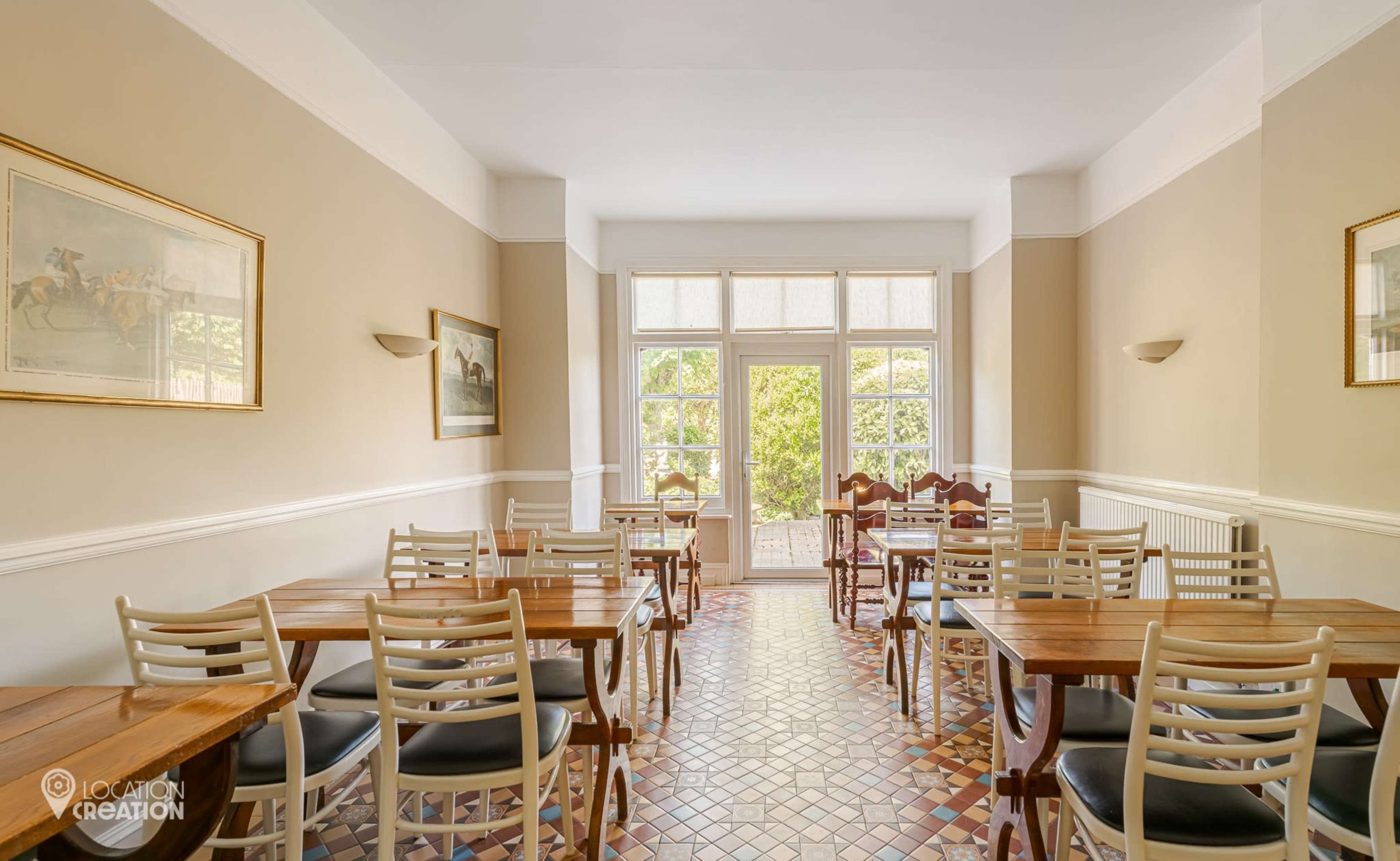 The image shows a dining area with wooden tables and chairs, featuring large windows that let in natural light, and tiled flooring.