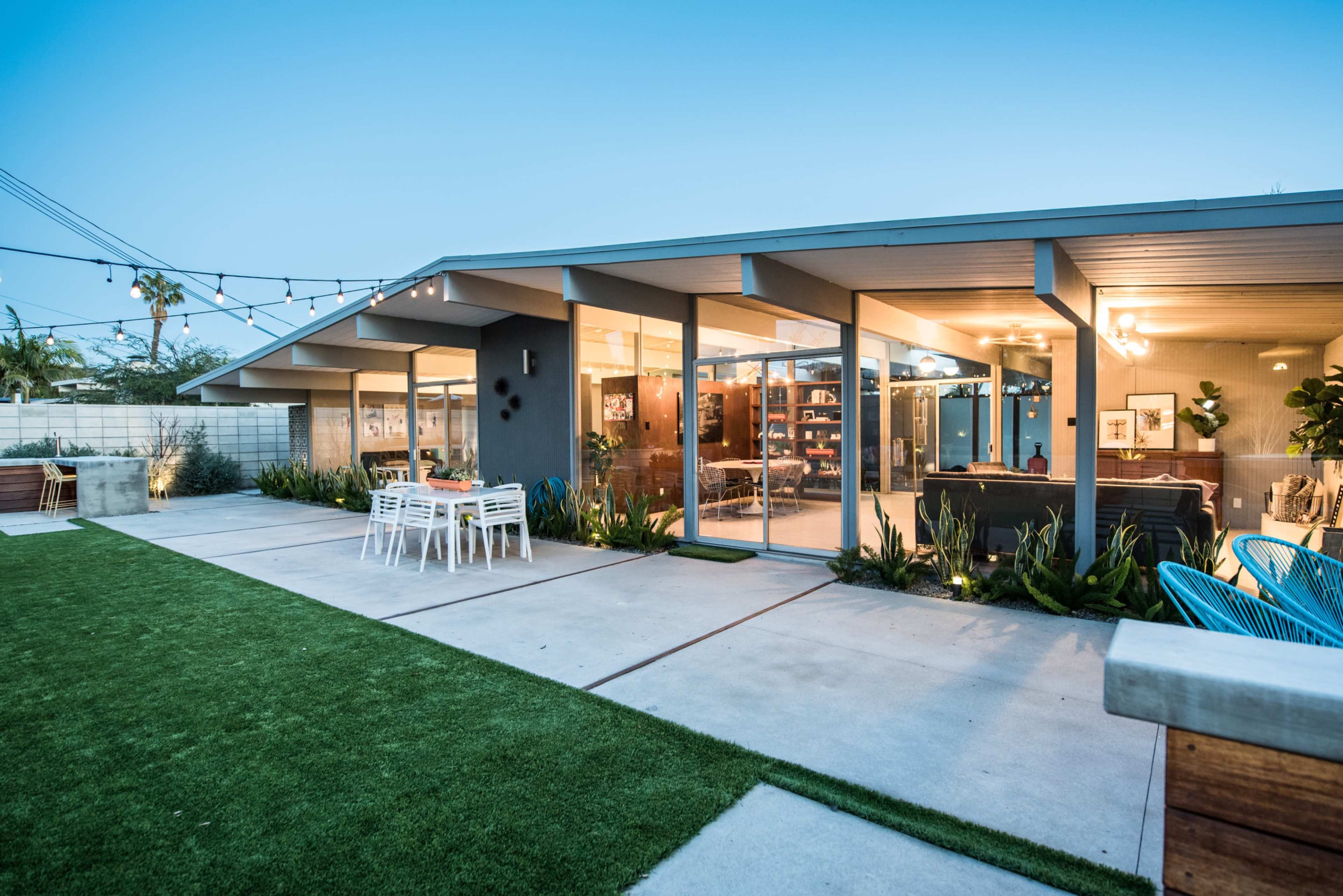 A modern single-story house features large glass windows and sliding doors facing a landscaped yard with patio furniture and outdoor lighting.