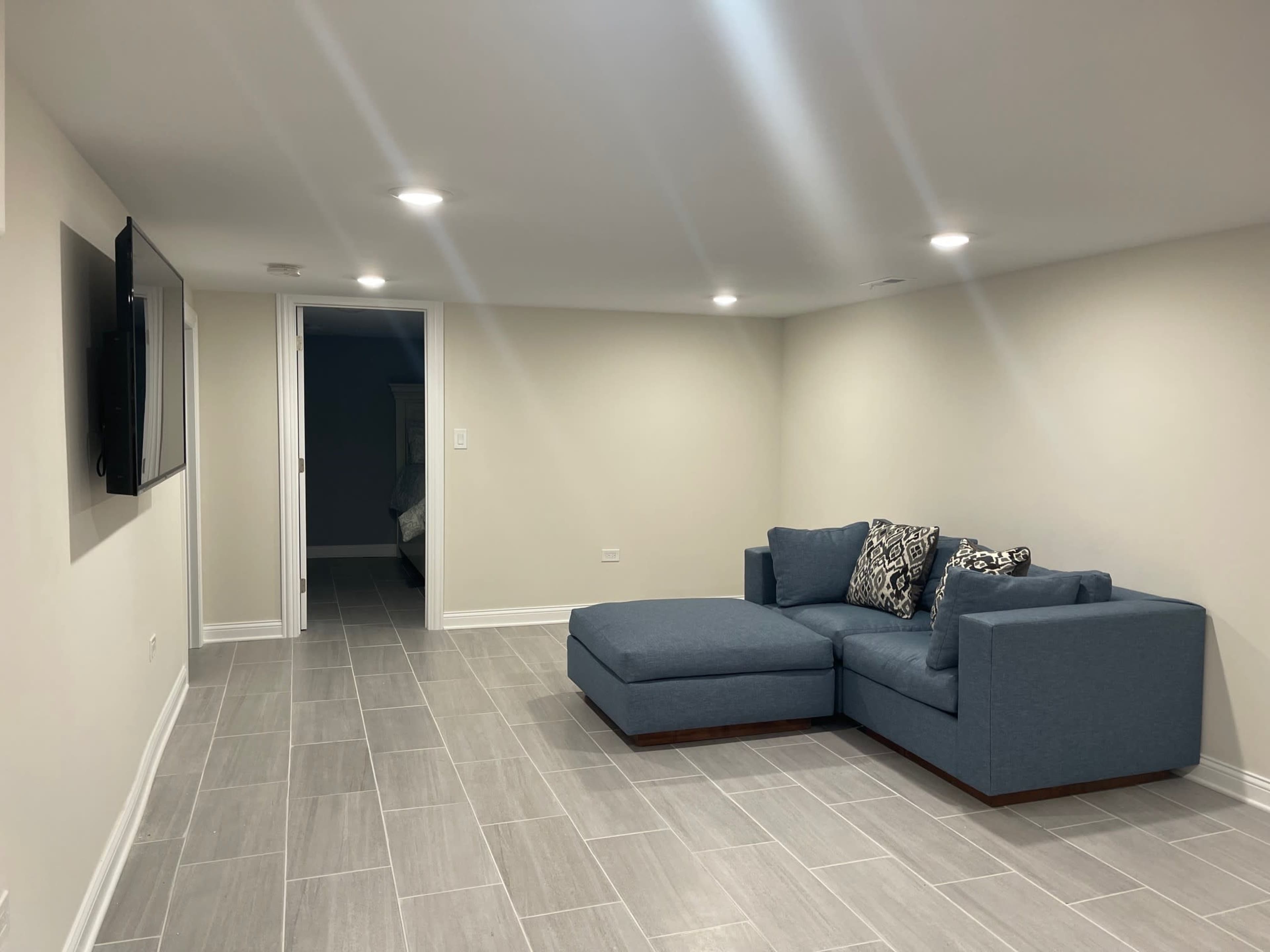 A finished basement with gray tile flooring, a blue sectional couch, and a wall-mounted television.