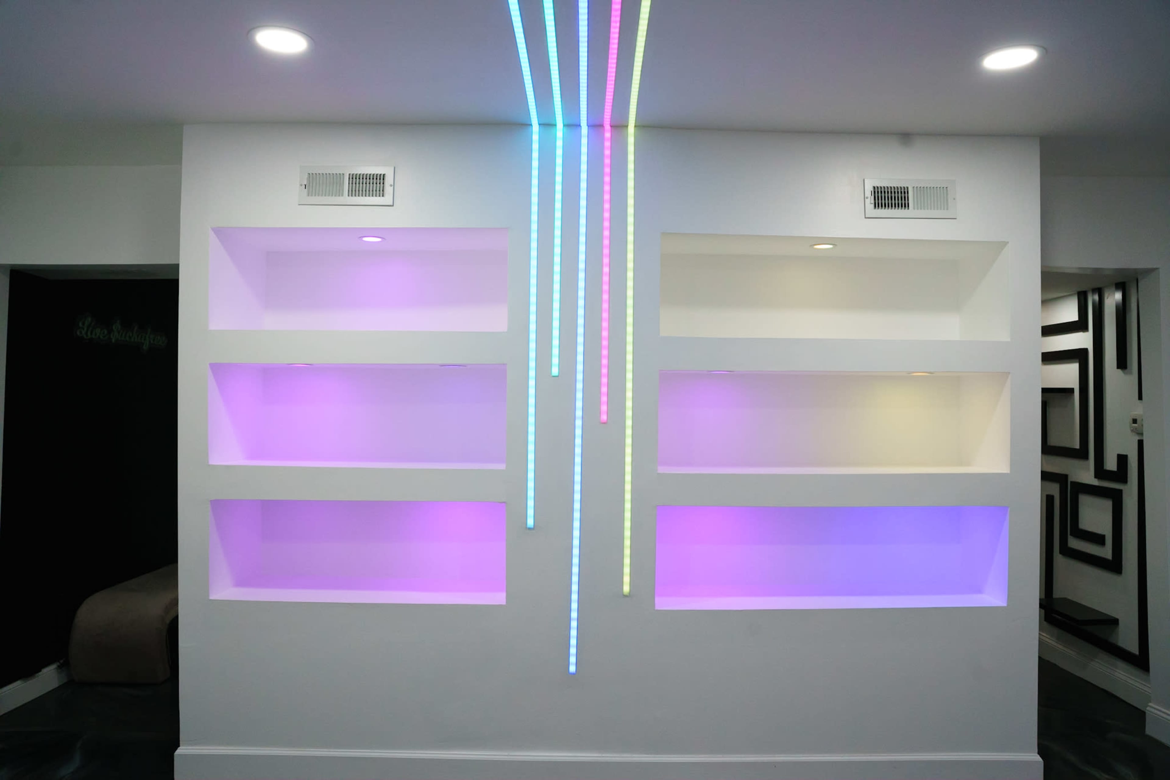 A wall features illuminated shelves with pink and blue lighting and three vertical light strips cascading downward.