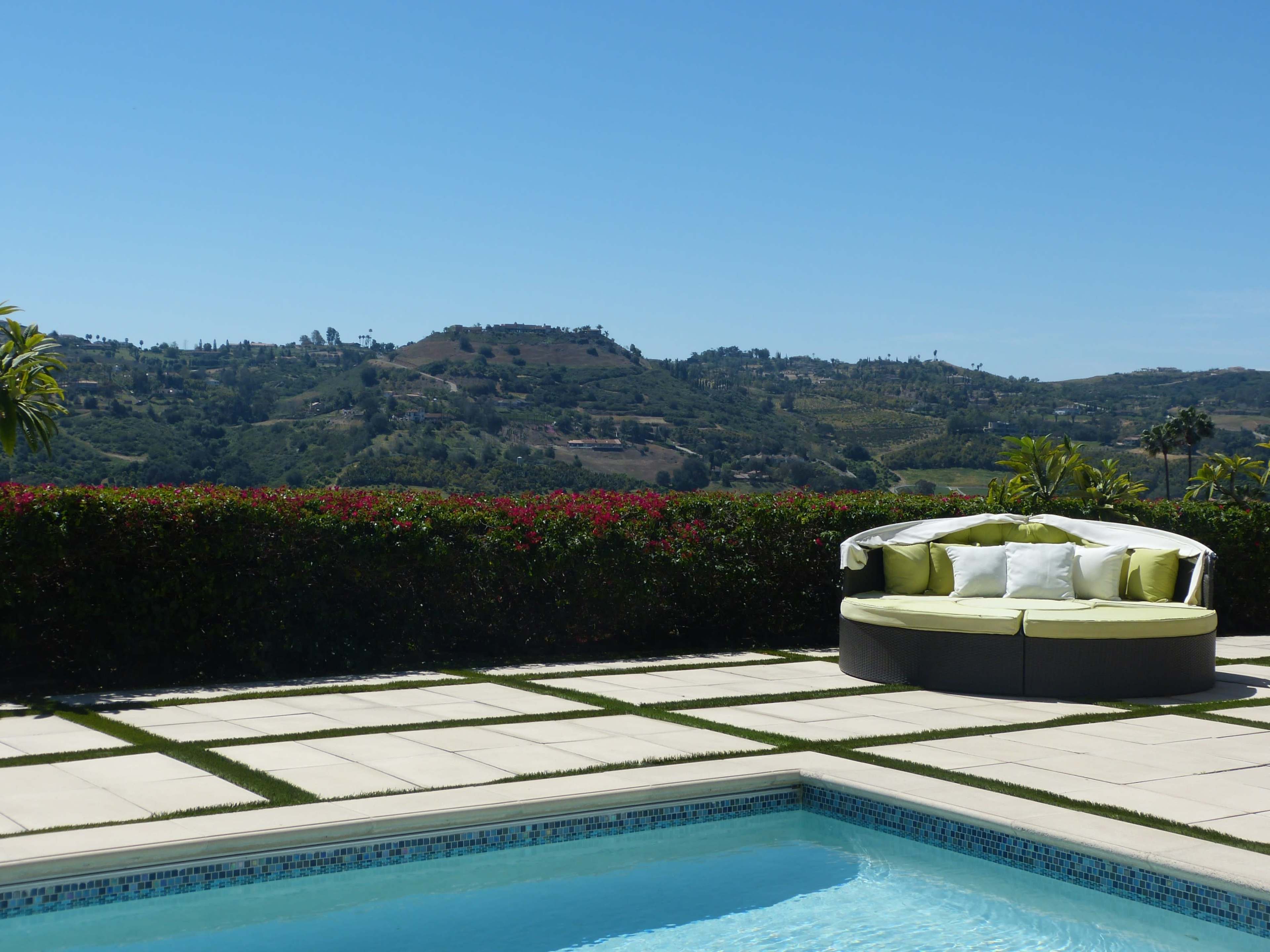 A circular seating area with green cushions surrounds a pool, set against a backdrop of hills and clear blue sky.