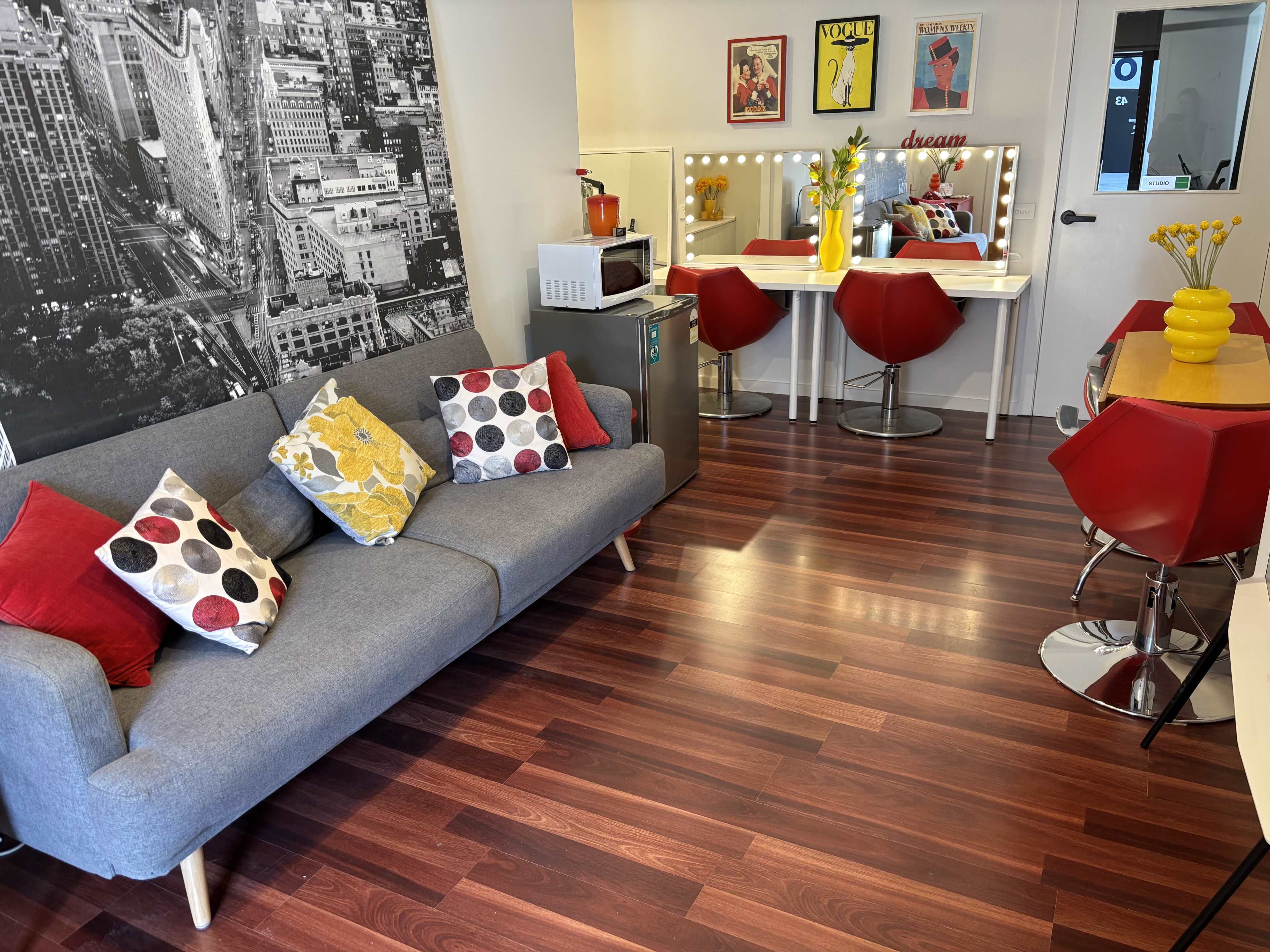 The image shows a modern living area featuring a gray sofa with colorful cushions, a small dining table with red chairs, and a vanity area illuminated by lights.