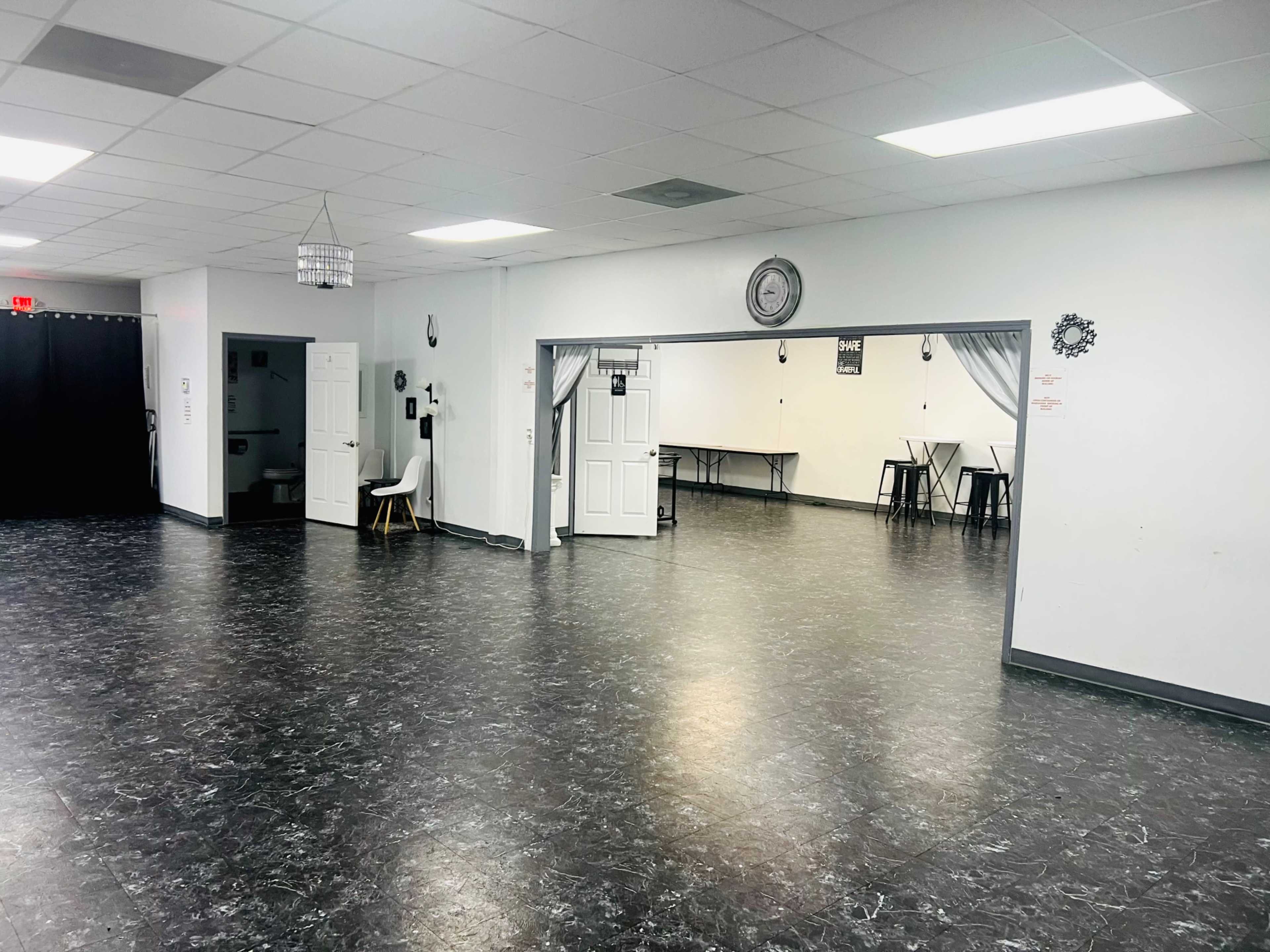 The image shows a spacious, empty room with dark flooring, white walls, and a doorway leading to another area with tables and chairs set up.