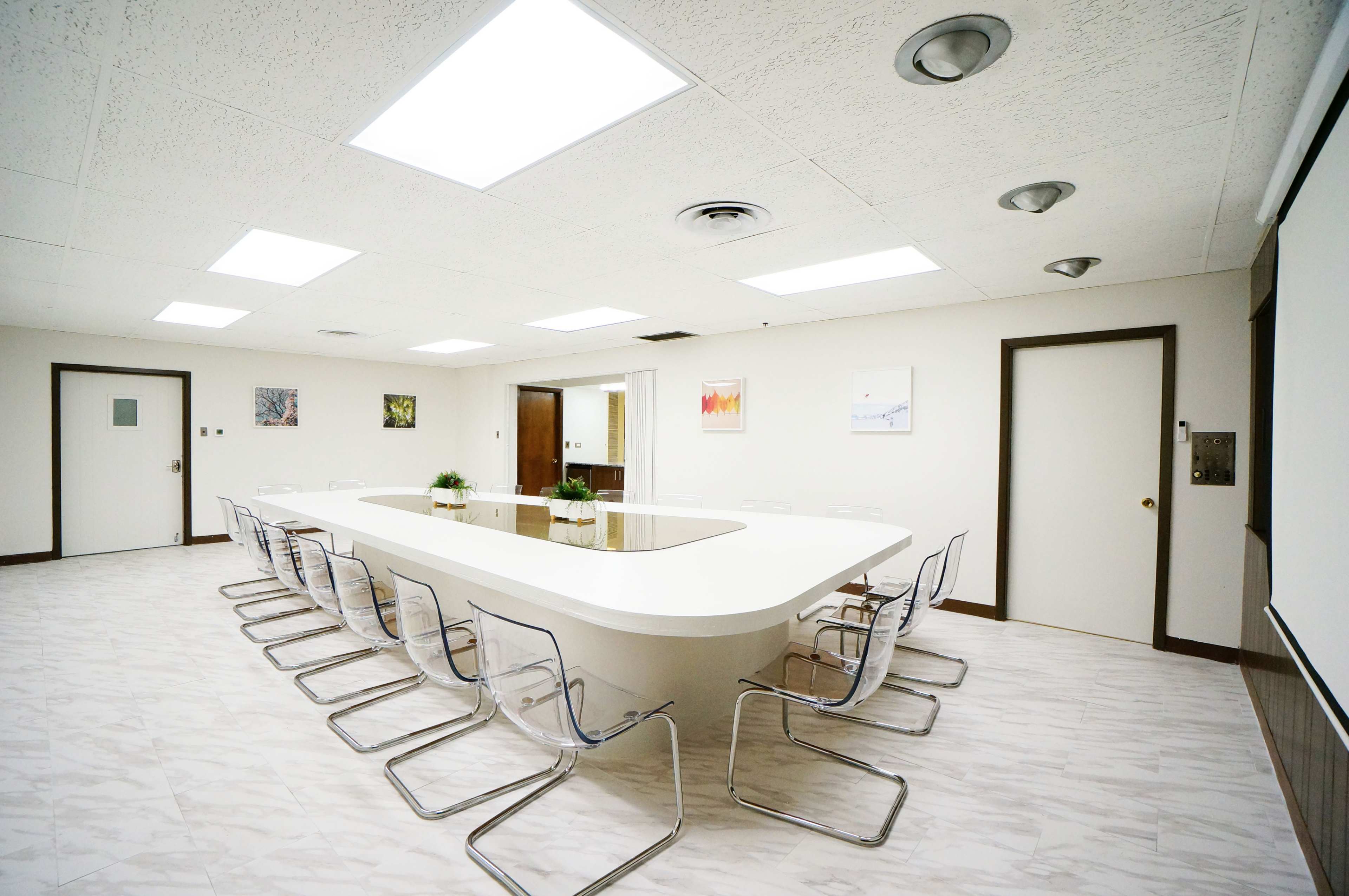 A modern conference room features a long white table surrounded by transparent chairs, with light fixtures illuminating the space.