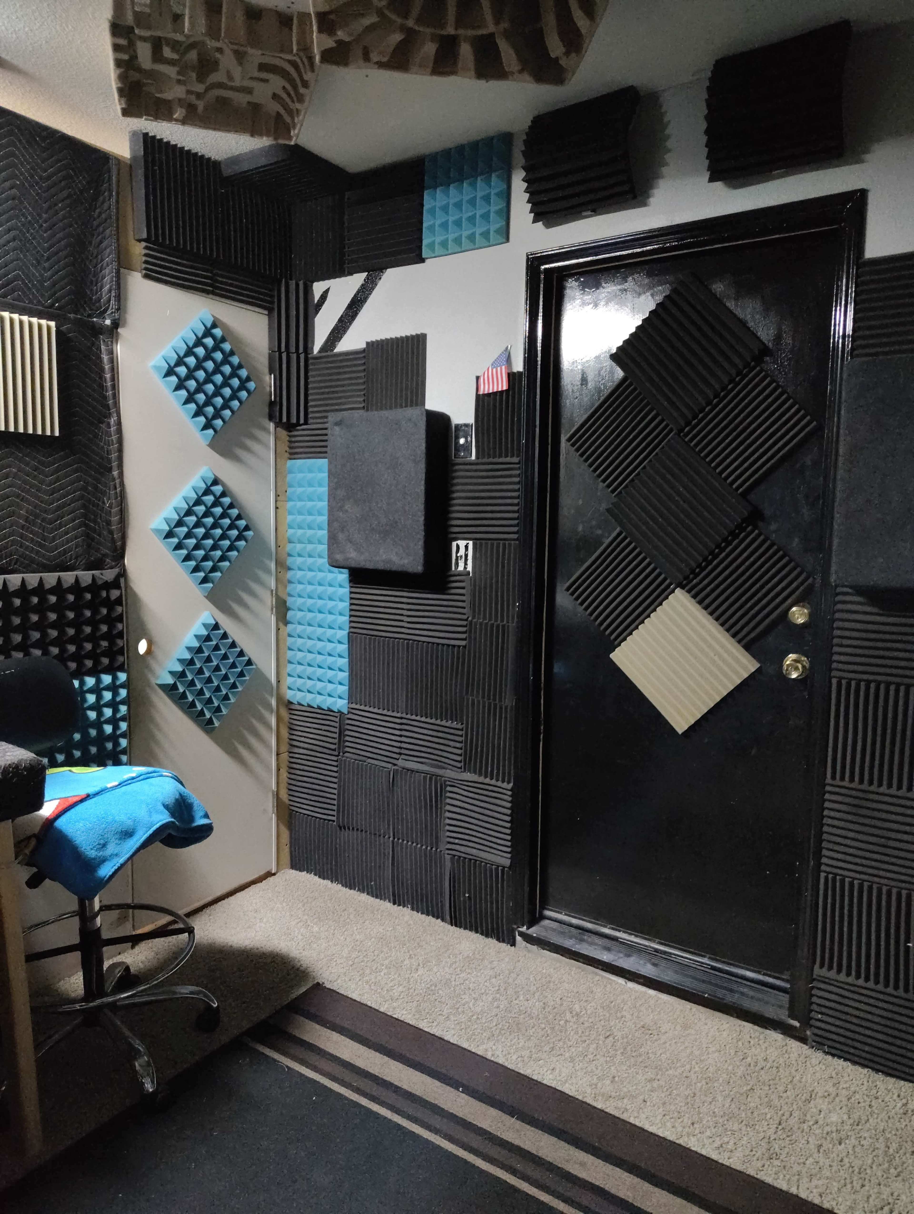 The image shows a soundproofed room with walls covered in various acoustic foam panels, a black door, and a swivel chair in the corner.