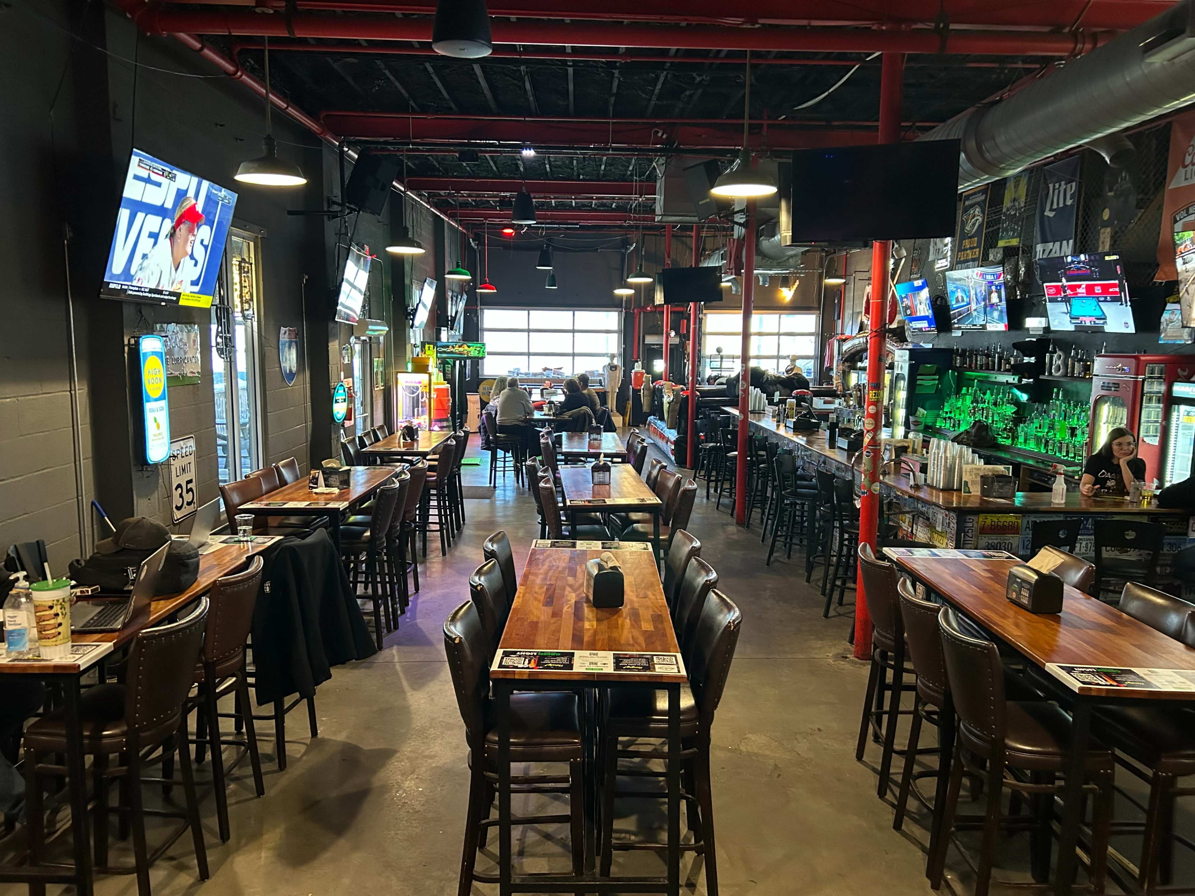 The image shows a lively sports bar with wooden tables, high-backed chairs, multiple televisions displaying sports, and a well-stocked bar area in the background.