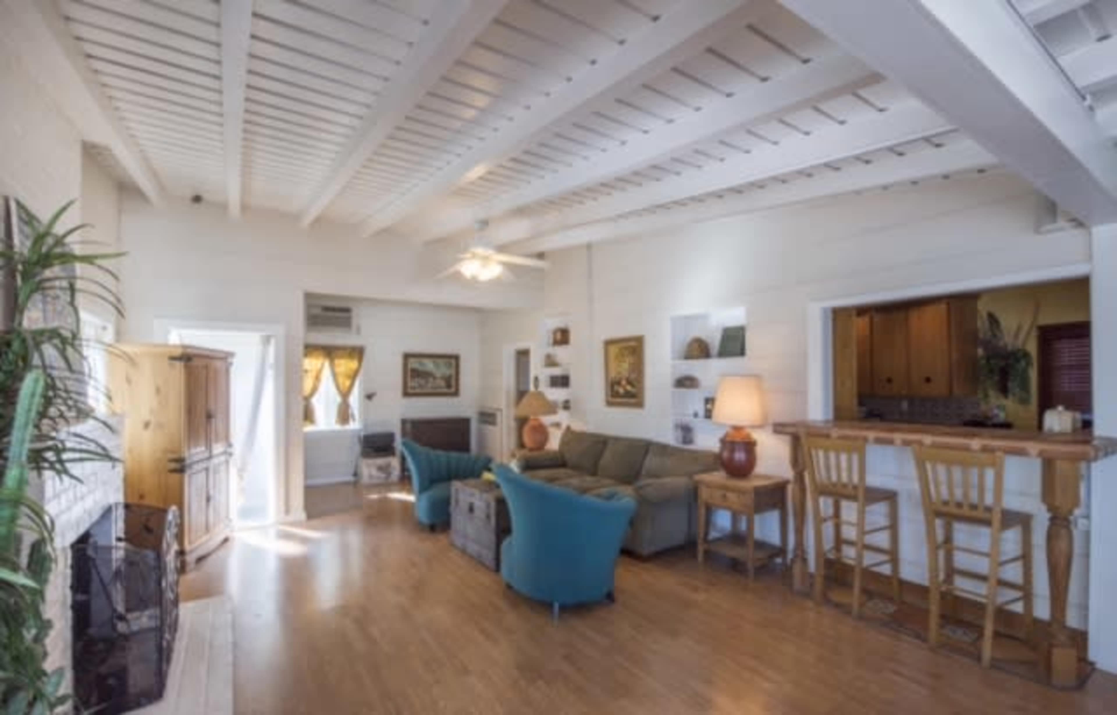 A bright living room with a light wooden floor, a green couch, a bar area with two stools, and a ceiling fan above.