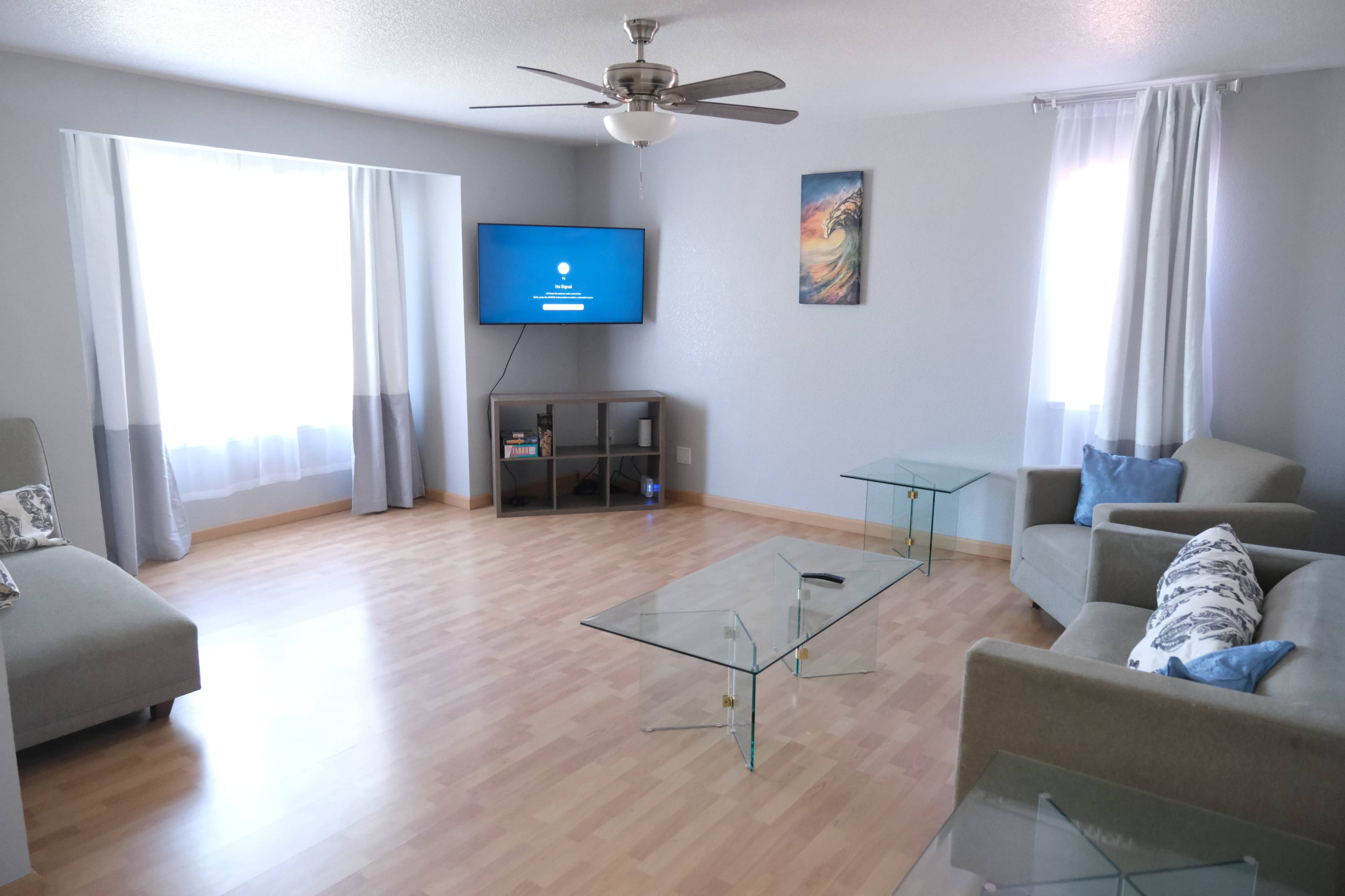 The image shows a light-colored living room with two sofas, a glass coffee table, a television mounted on the wall, and large windows draped with sheer curtains.
