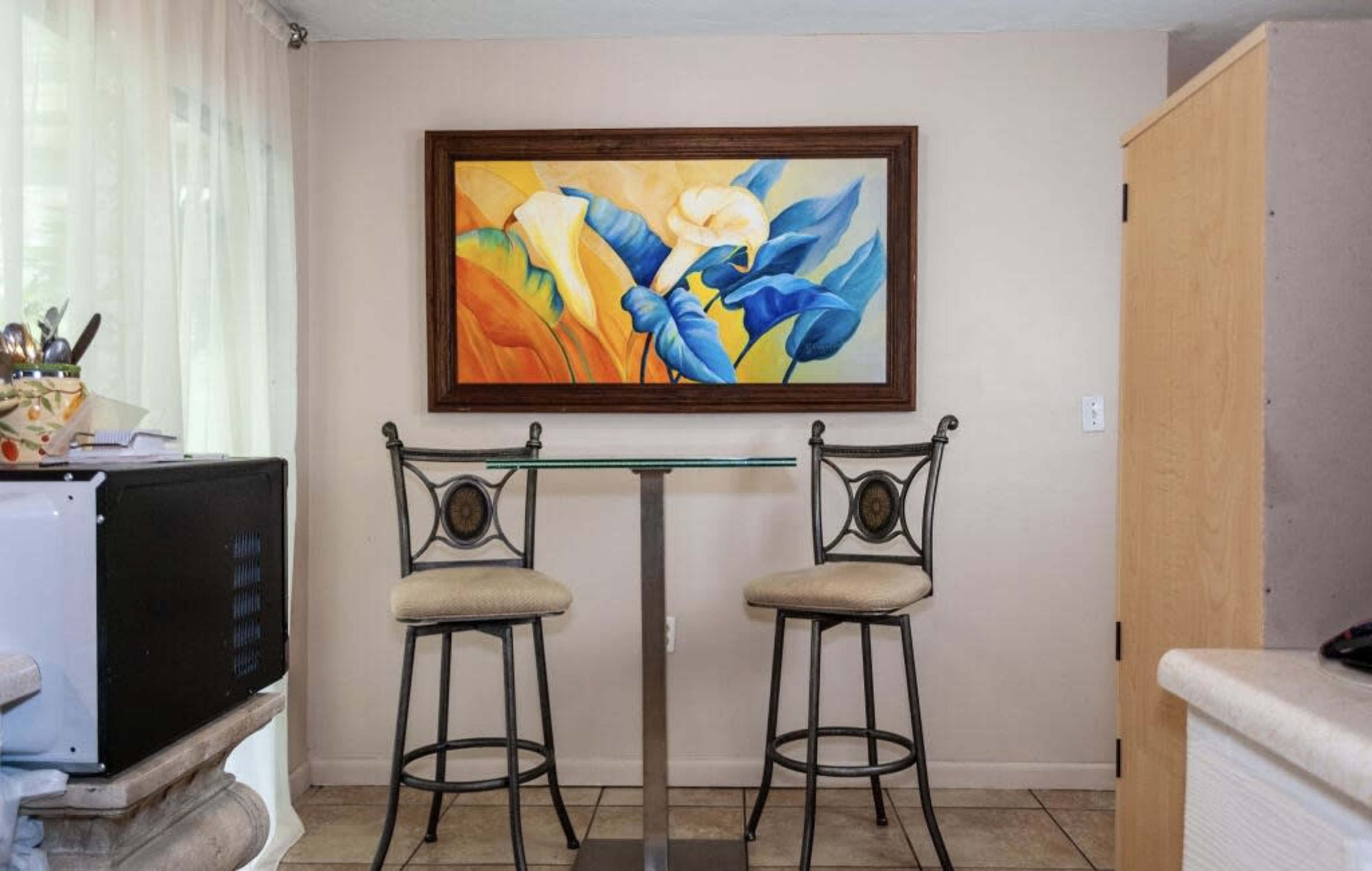 A small dining area with two metal bar stools at a glass table, accompanied by a colorful framed painting on the wall.