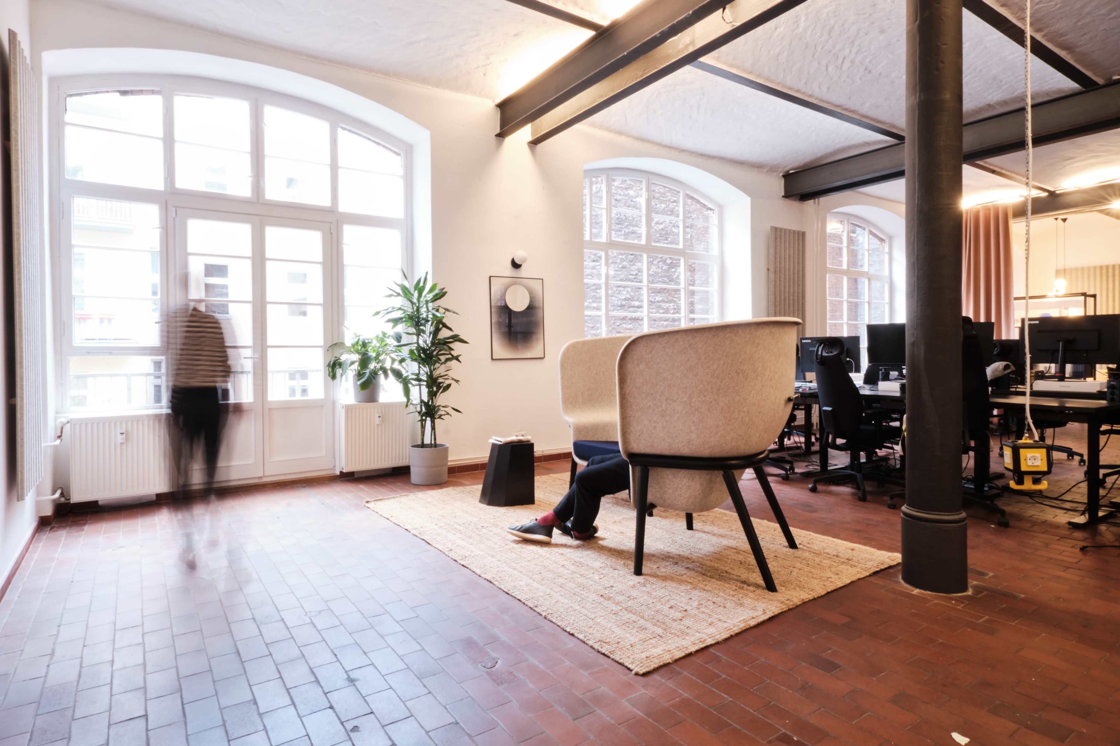The image shows a spacious office interior with a large window, a decorative plant, and a seating area featuring two armchairs on a woven rug, alongside workstations in the background.