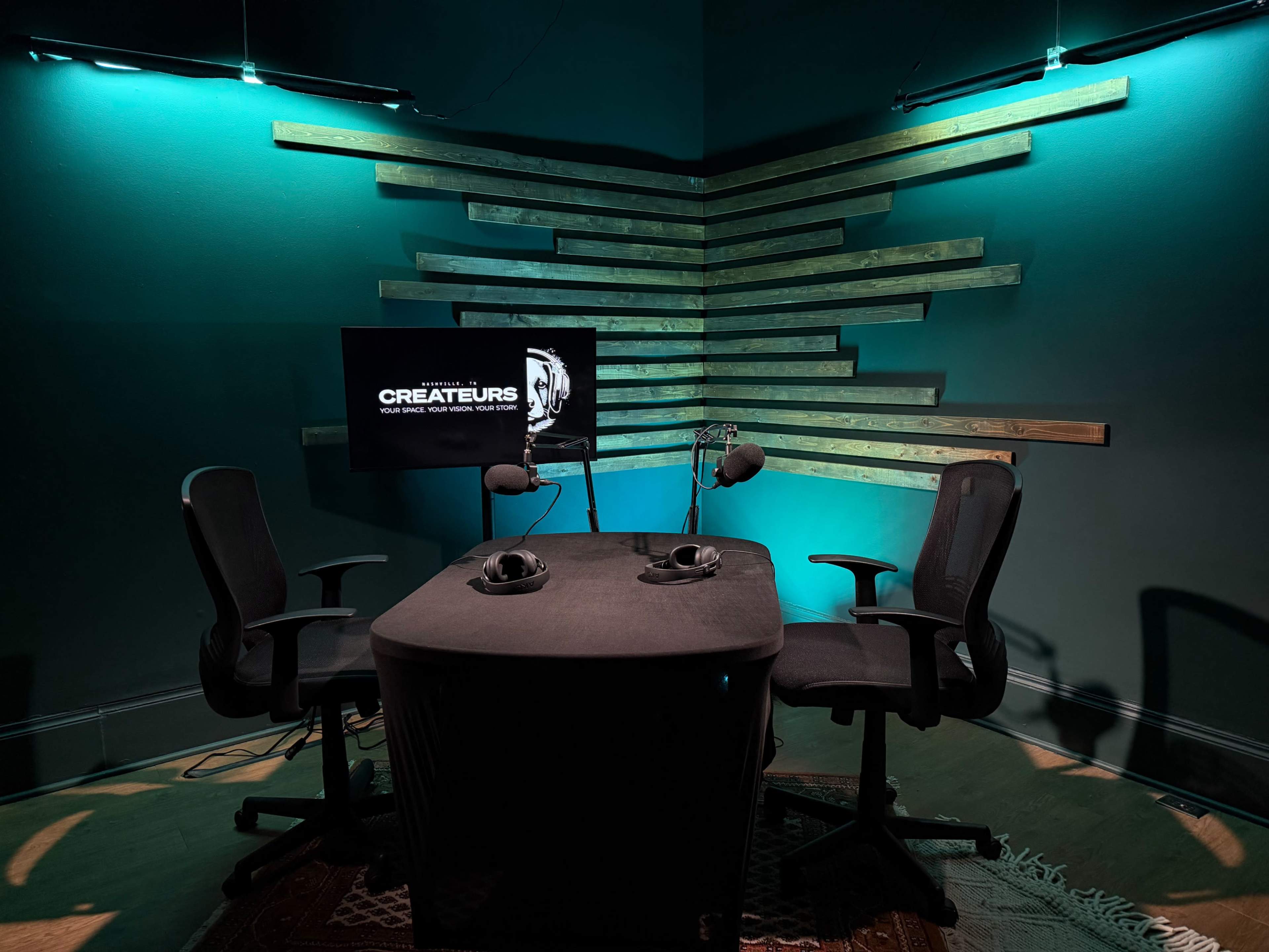 The image shows a podcast studio featuring a dark table, two black chairs, and a screen displaying the word "CREATEURS" against a backdrop of teal and wooden accents.