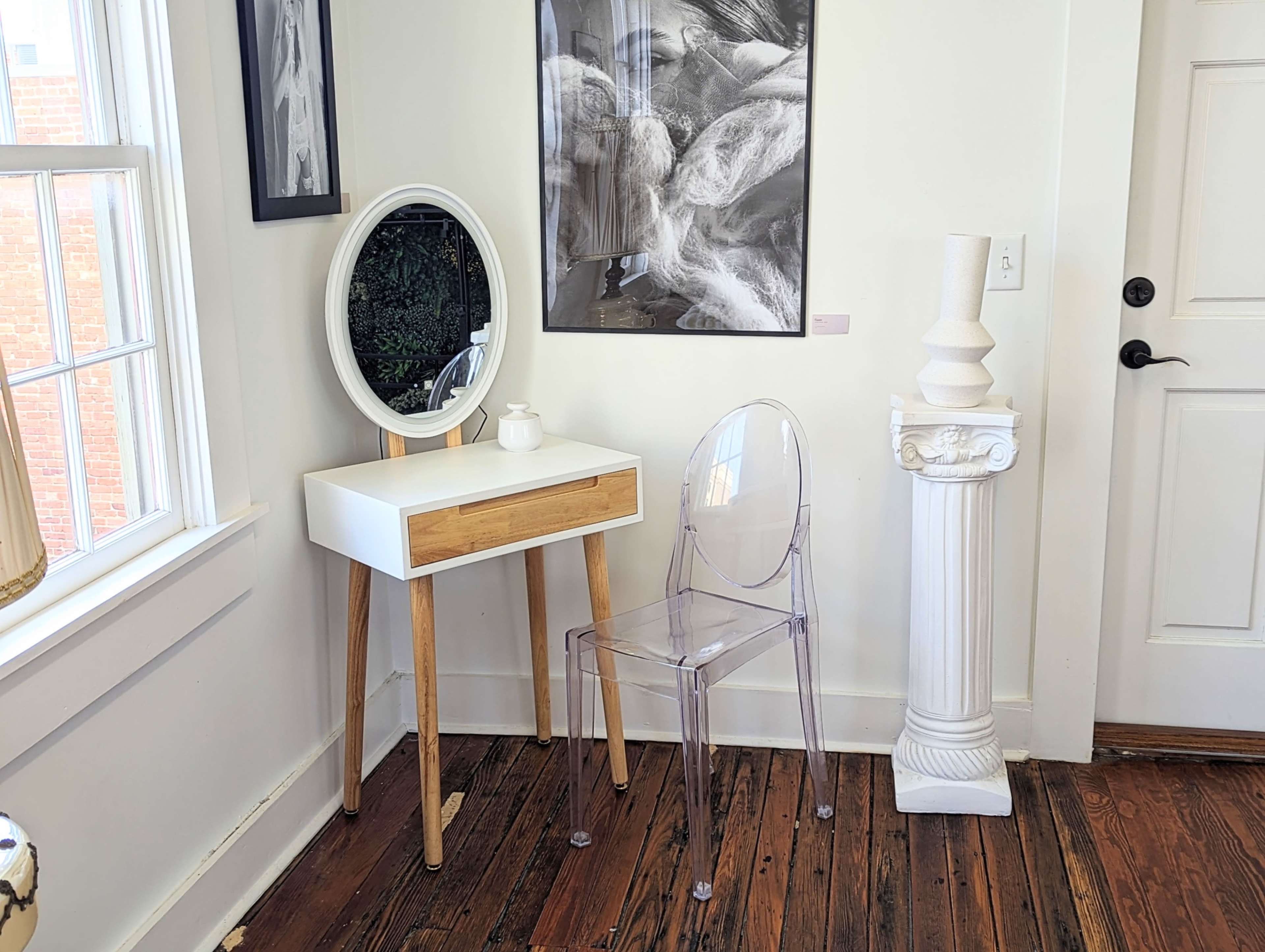 A minimalist corner of a room featuring a white and wood vanity table with a round mirror, a clear plastic chair, and a decorative column beside it.