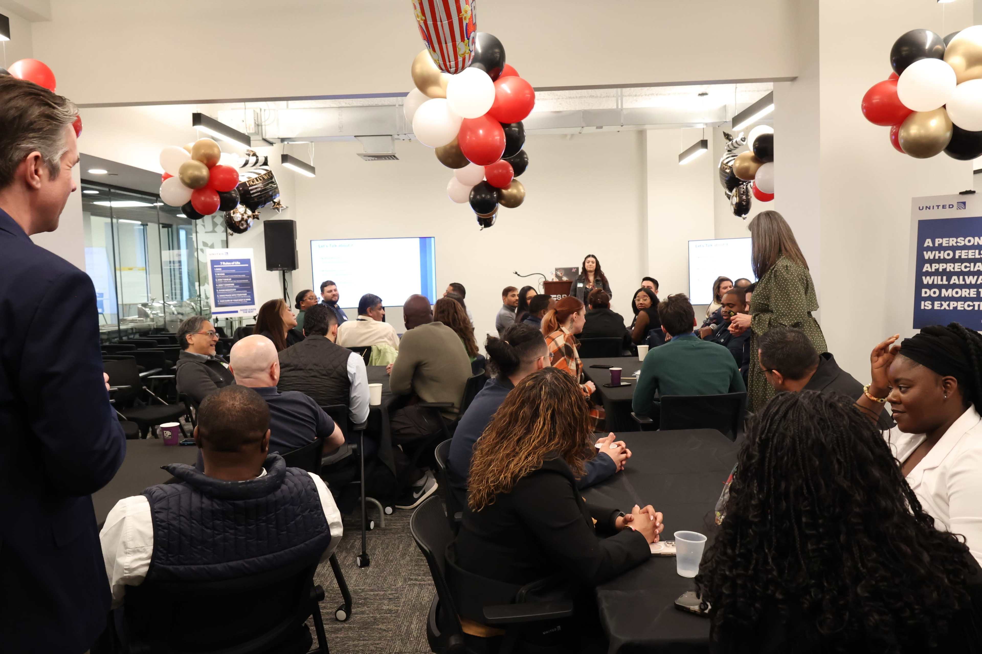 A group of diverse individuals gathers around tables inside a conference room, listening to a speaker in front of a backdrop decorated with balloons.