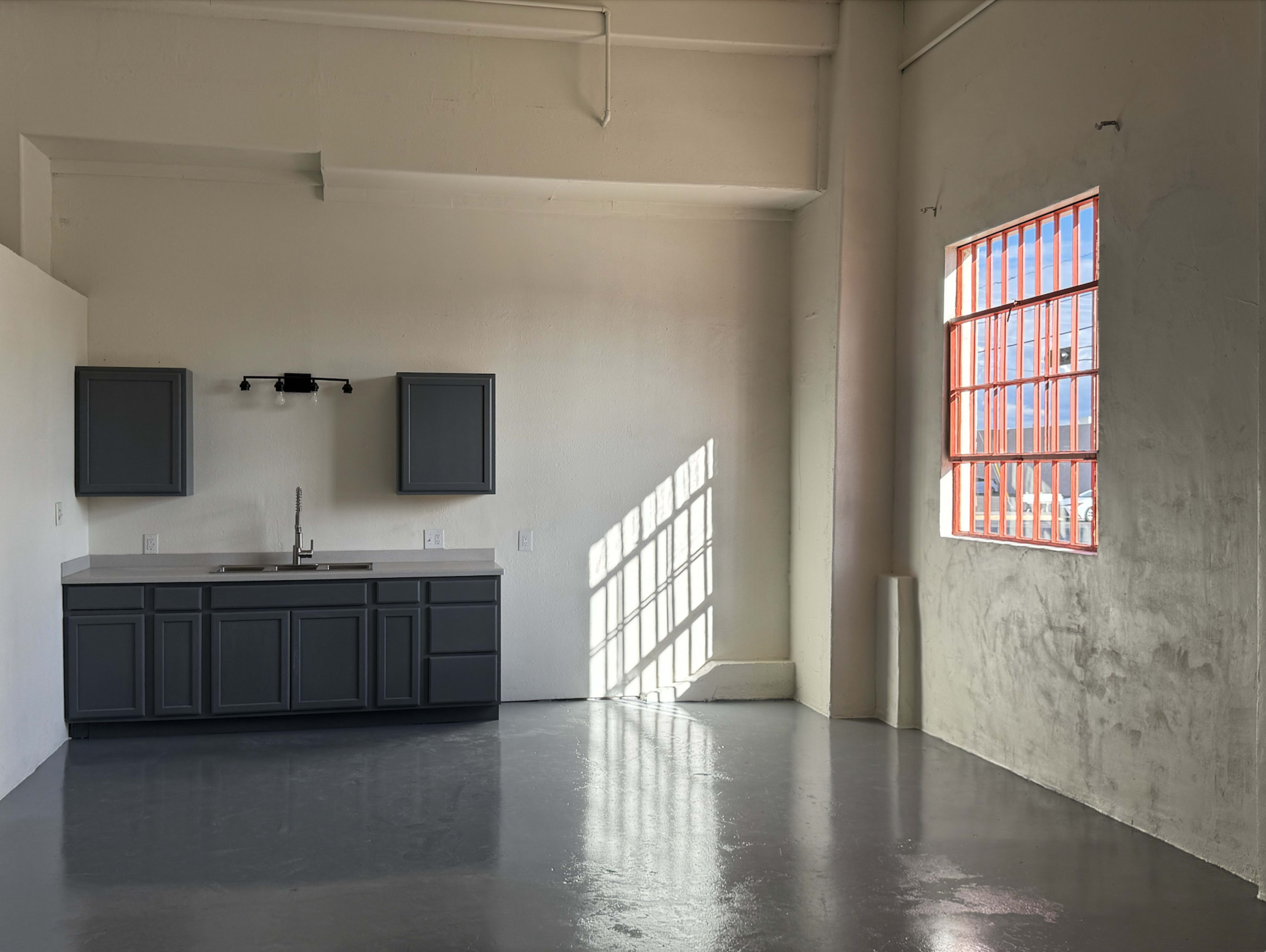 The image shows an empty, minimally furnished room with a kitchen area featuring dark cabinets and a large window with red framing that casts shadows on the floor.