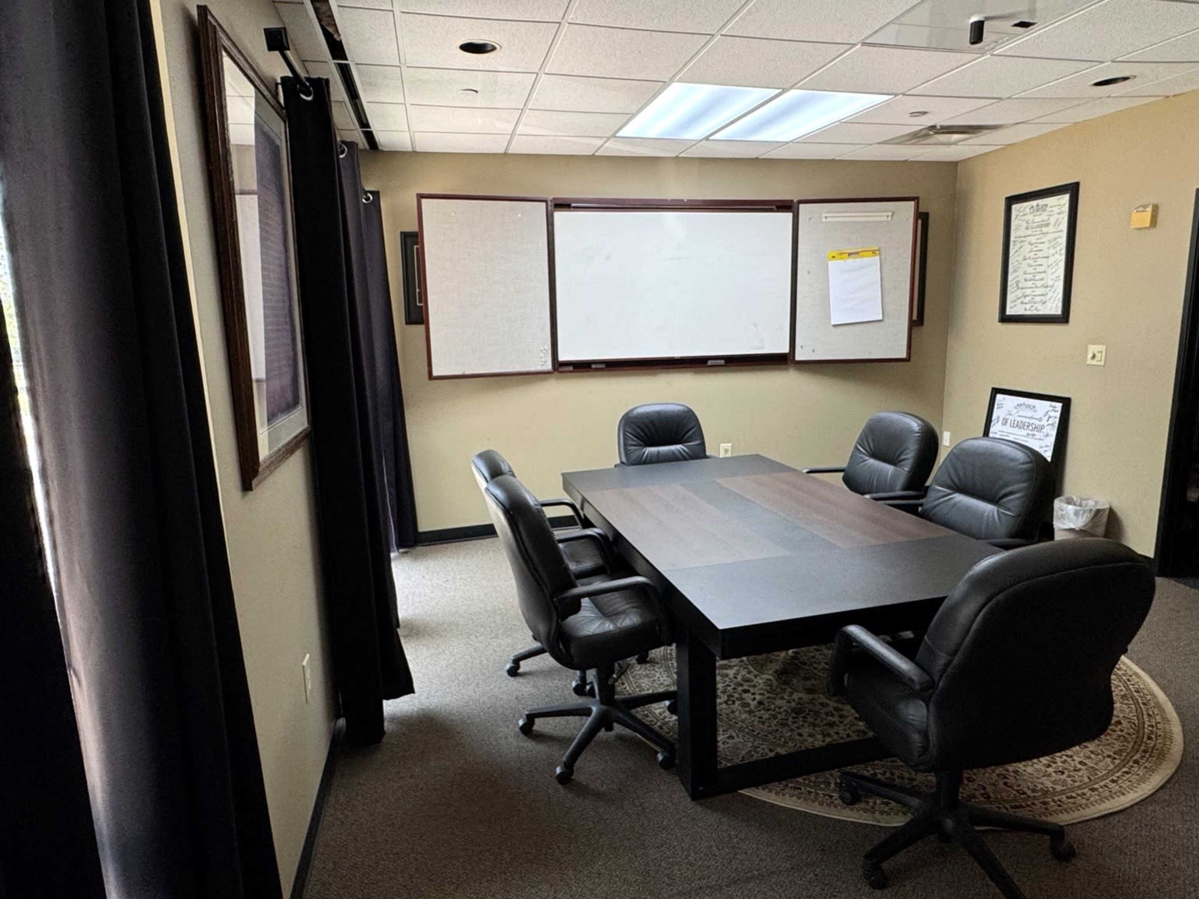 The image shows a conference room with a large table surrounded by black chairs, a whiteboard, and framed documents on the walls.