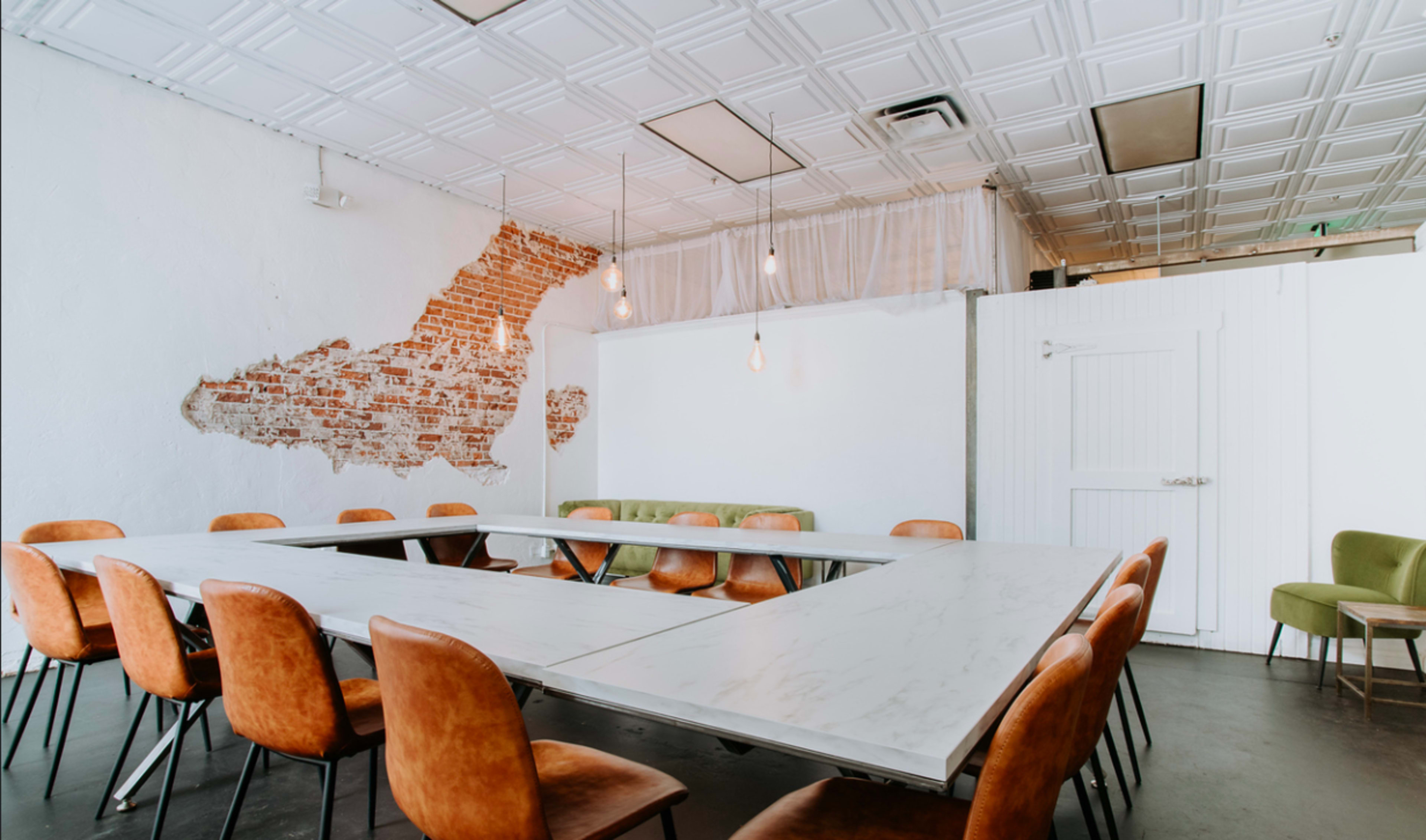 The image shows a modern meeting room with a long rectangular table surrounded by brown chairs, featuring exposed brick walls and pendant lighting.