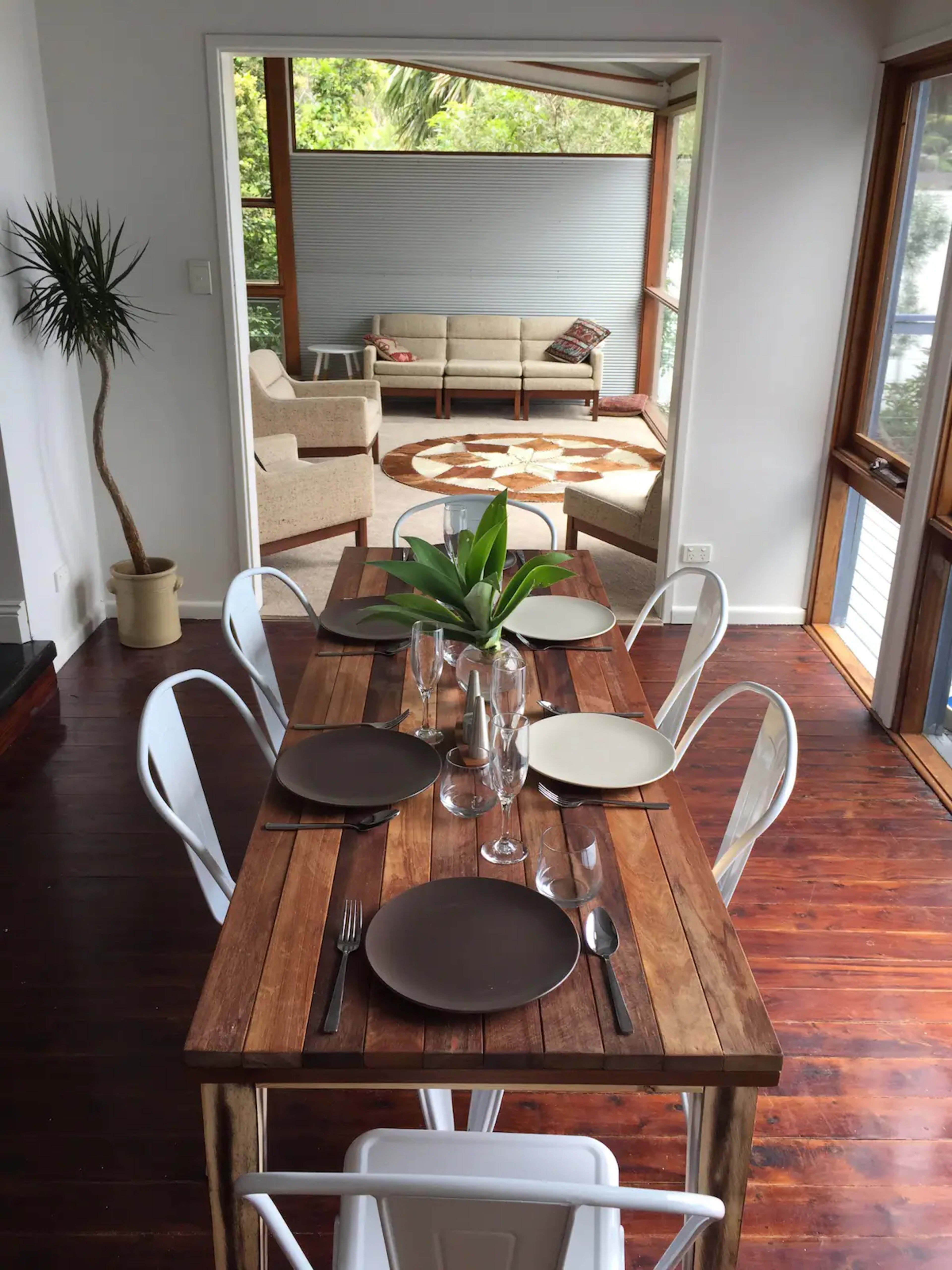 A wooden dining table is set with plates and glasses in a brightly lit room that opens to a cozy living area.