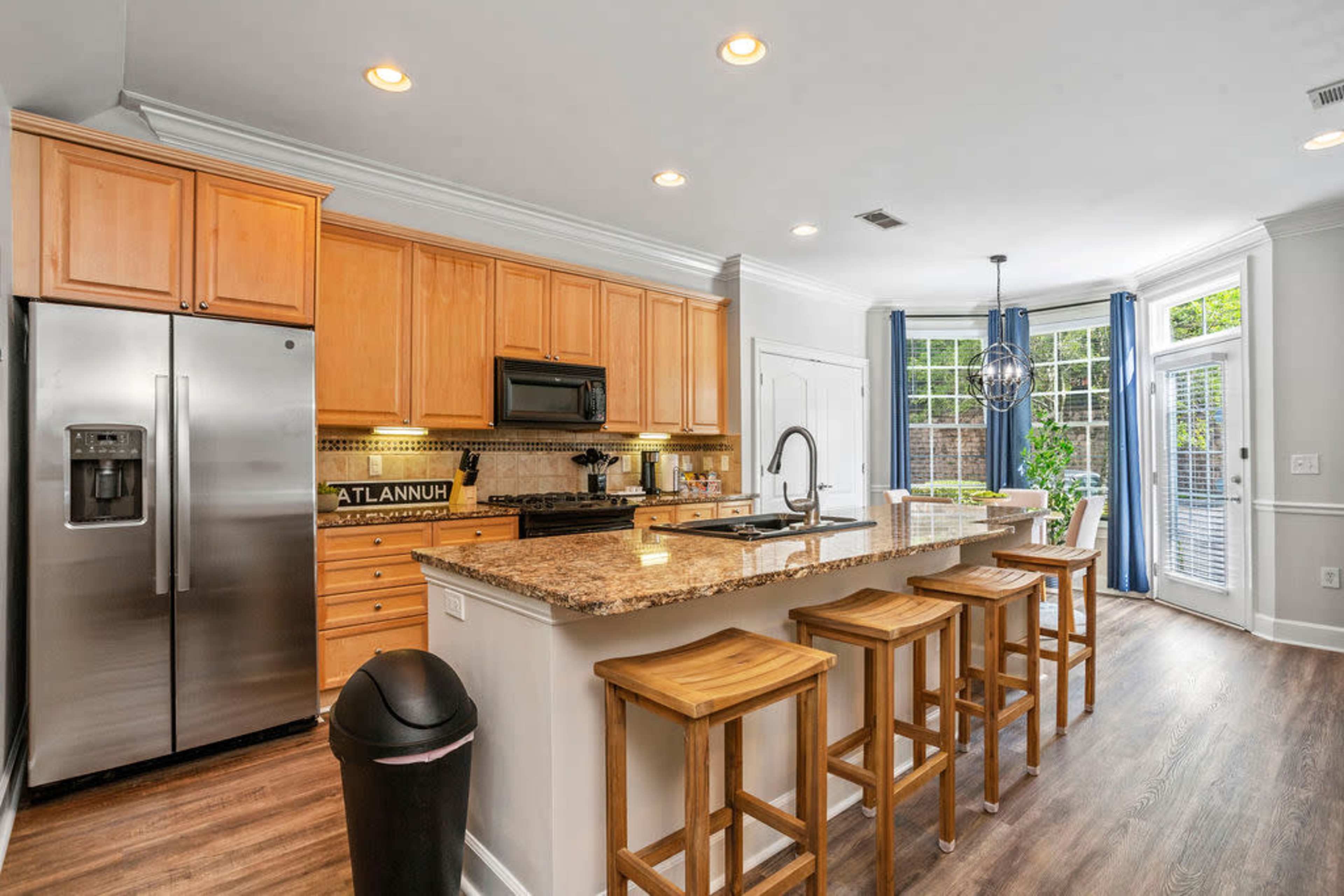 The image shows a modern kitchen with wooden cabinets, stainless steel appliances, a granite countertop, and seating for four at a kitchen island.