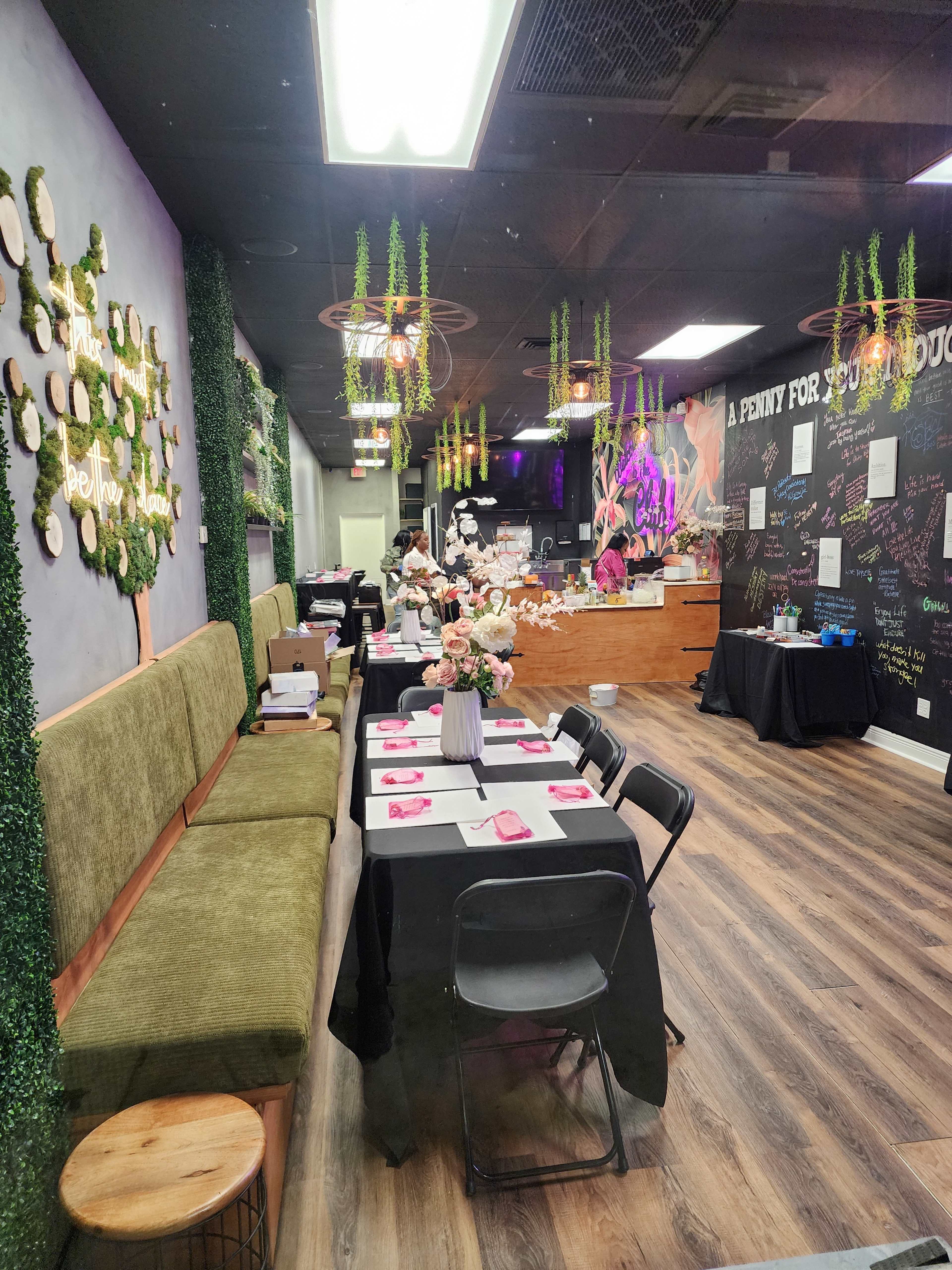 The image shows a cozy café-style interior with a long table set for an event, decorated with flowers and surrounded by greenery.