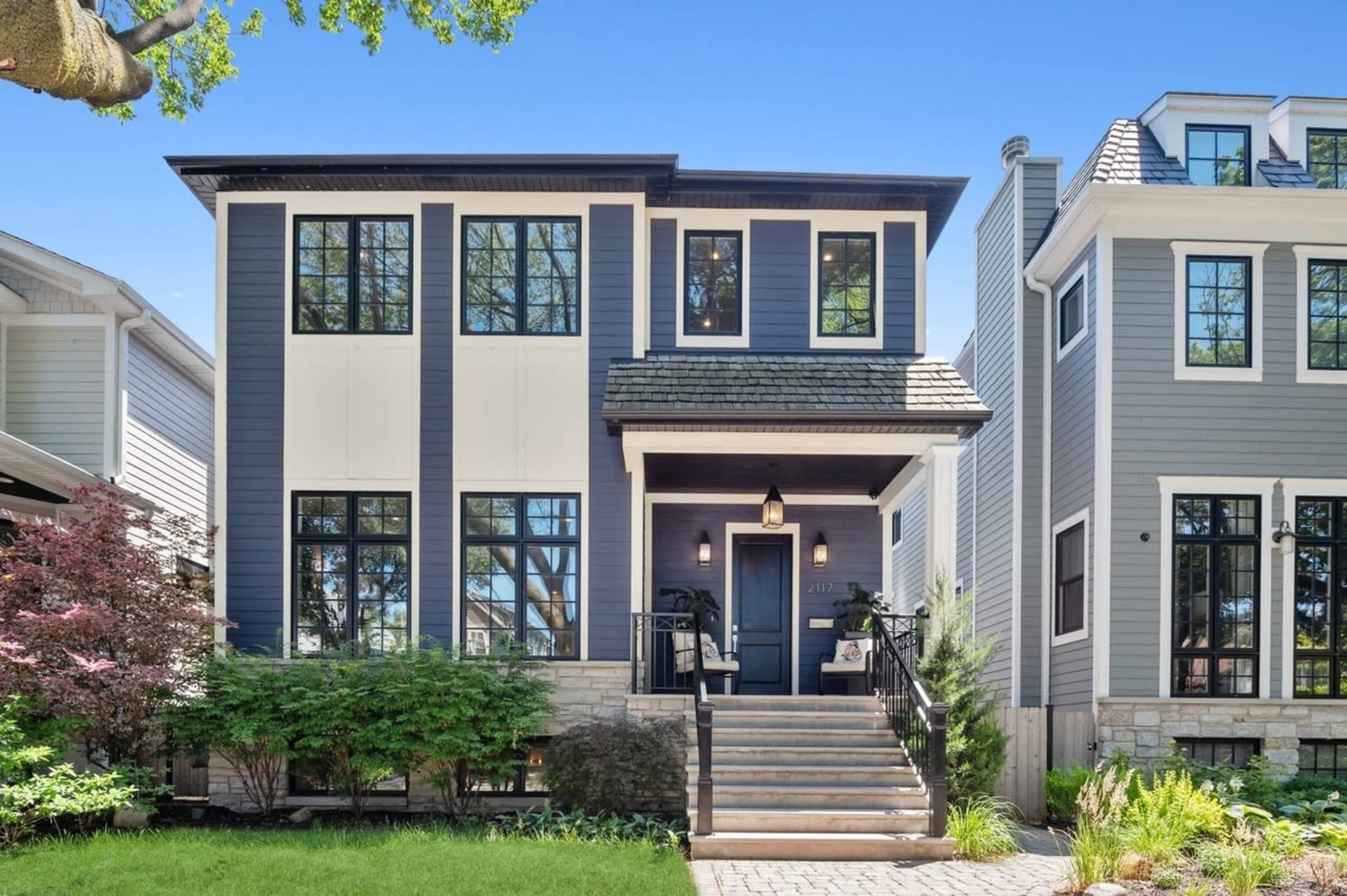 A modern two-story house with blue siding, large windows, and a stone walkway leads to its front steps.