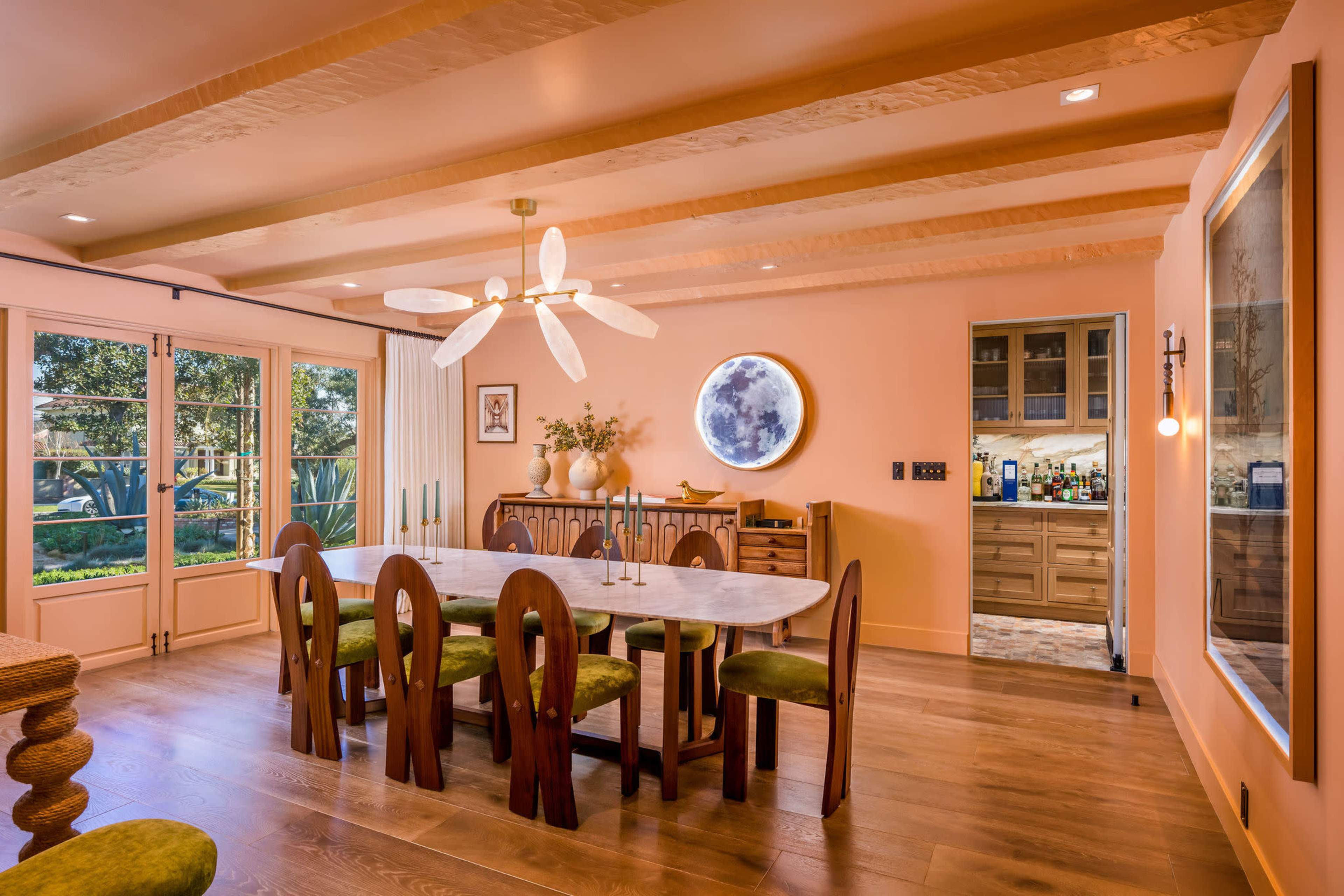 The image shows a modern dining room with a large marble table surrounded by wooden chairs and a round mirror on the wall, featuring a view of a landscaped garden through large windows.