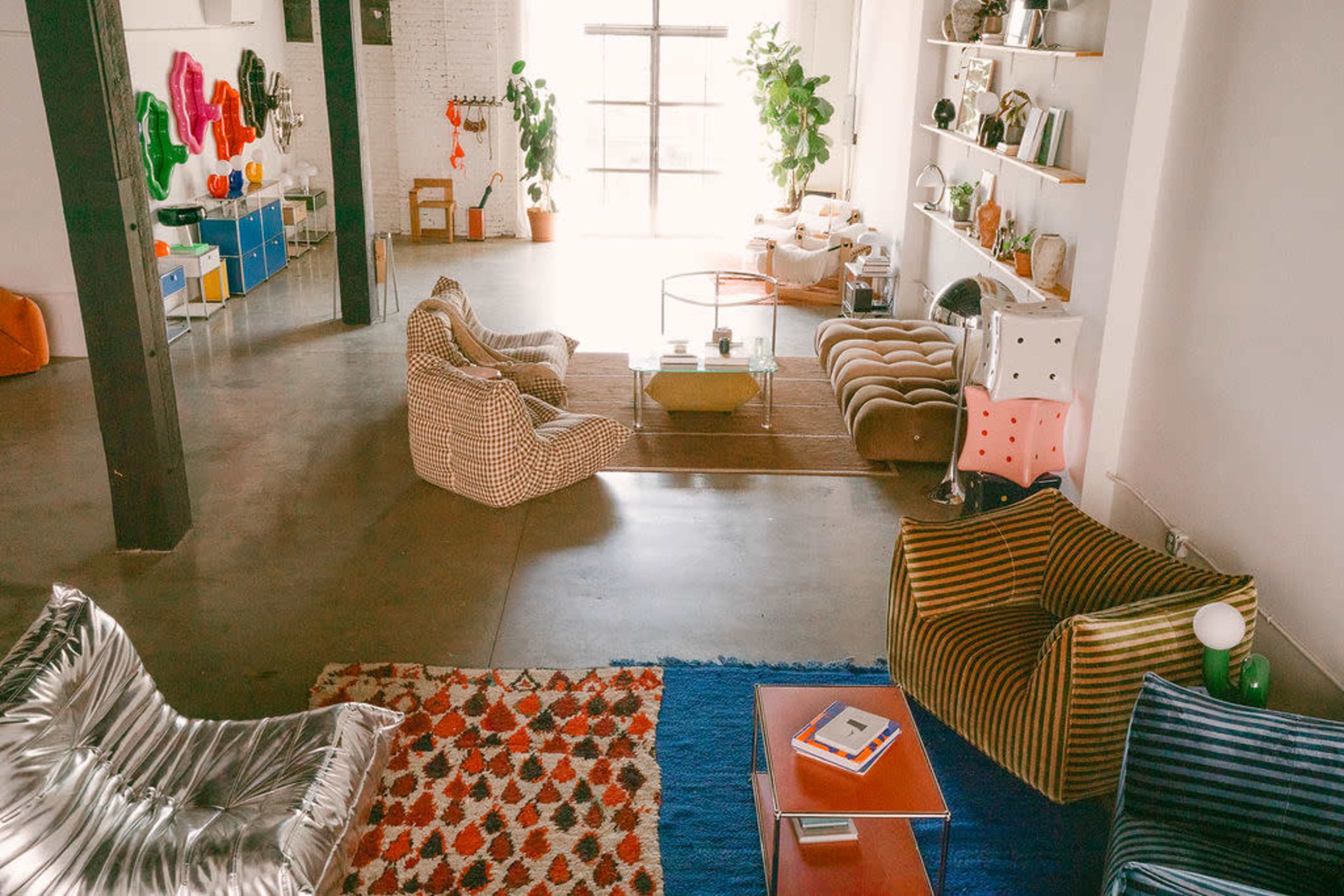 The image shows a spacious, modern living area with various styles of seating, colorful decor, and large windows allowing natural light to filter in.