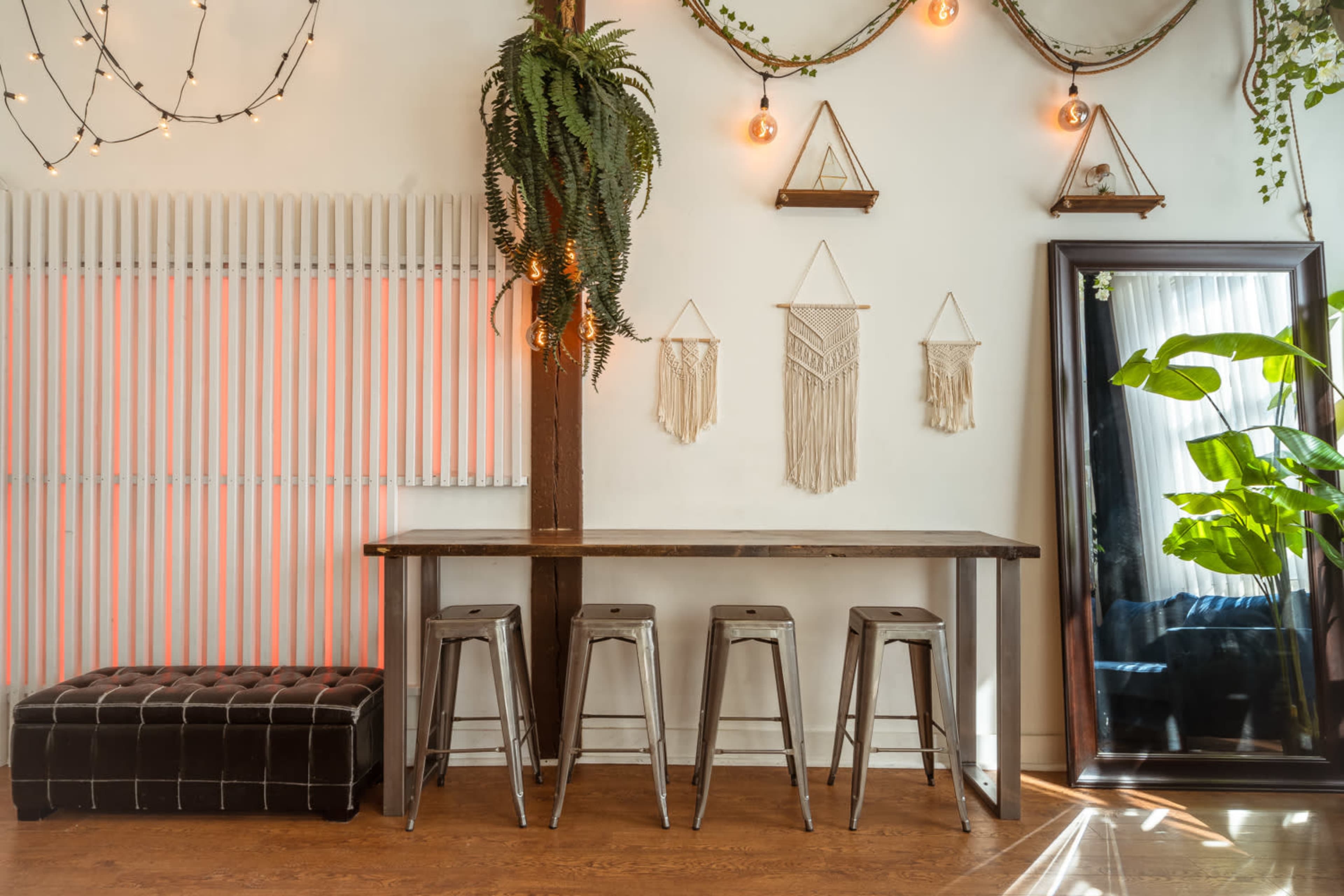 A wooden table with metal stools is set against a wall adorned with plants, macramé hangings, and illuminated light bulbs.