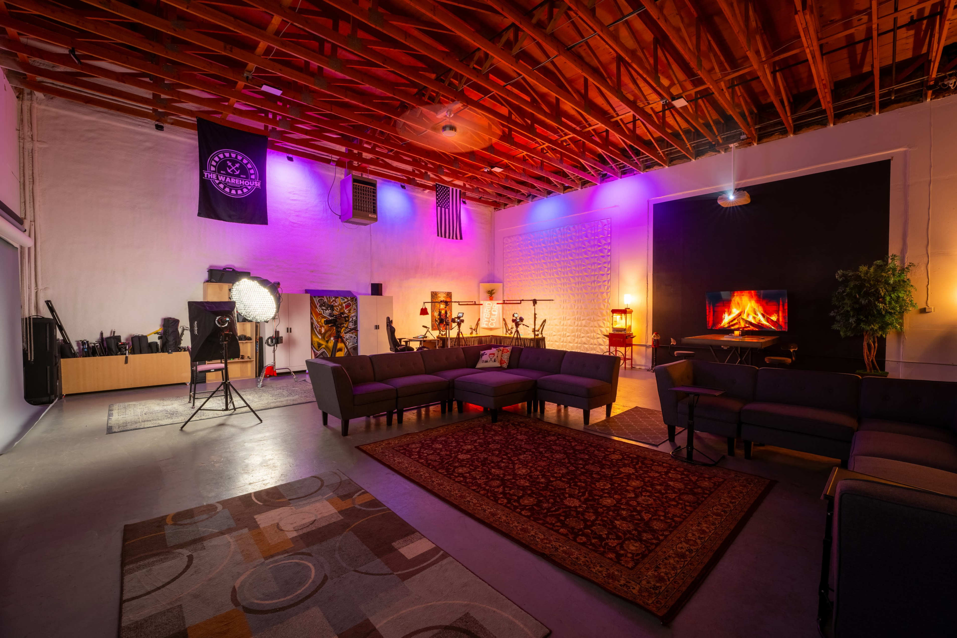 A spacious lounge area with couches, a decorative rug, and various lighting elements, featuring a faux fireplace and a backdrop of flags and musical instruments.