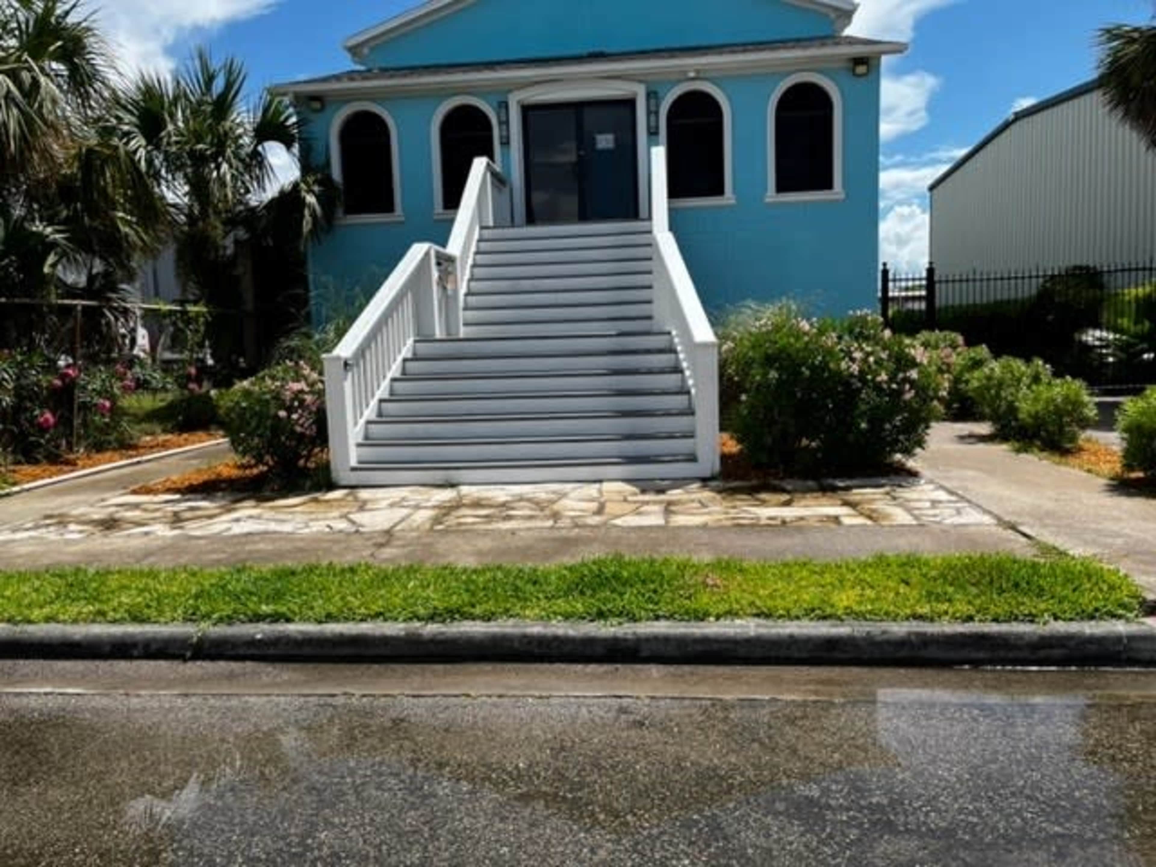A light blue building with a set of white stairs leading up to a double-door entrance, surrounded by greenery and a paved walkway.