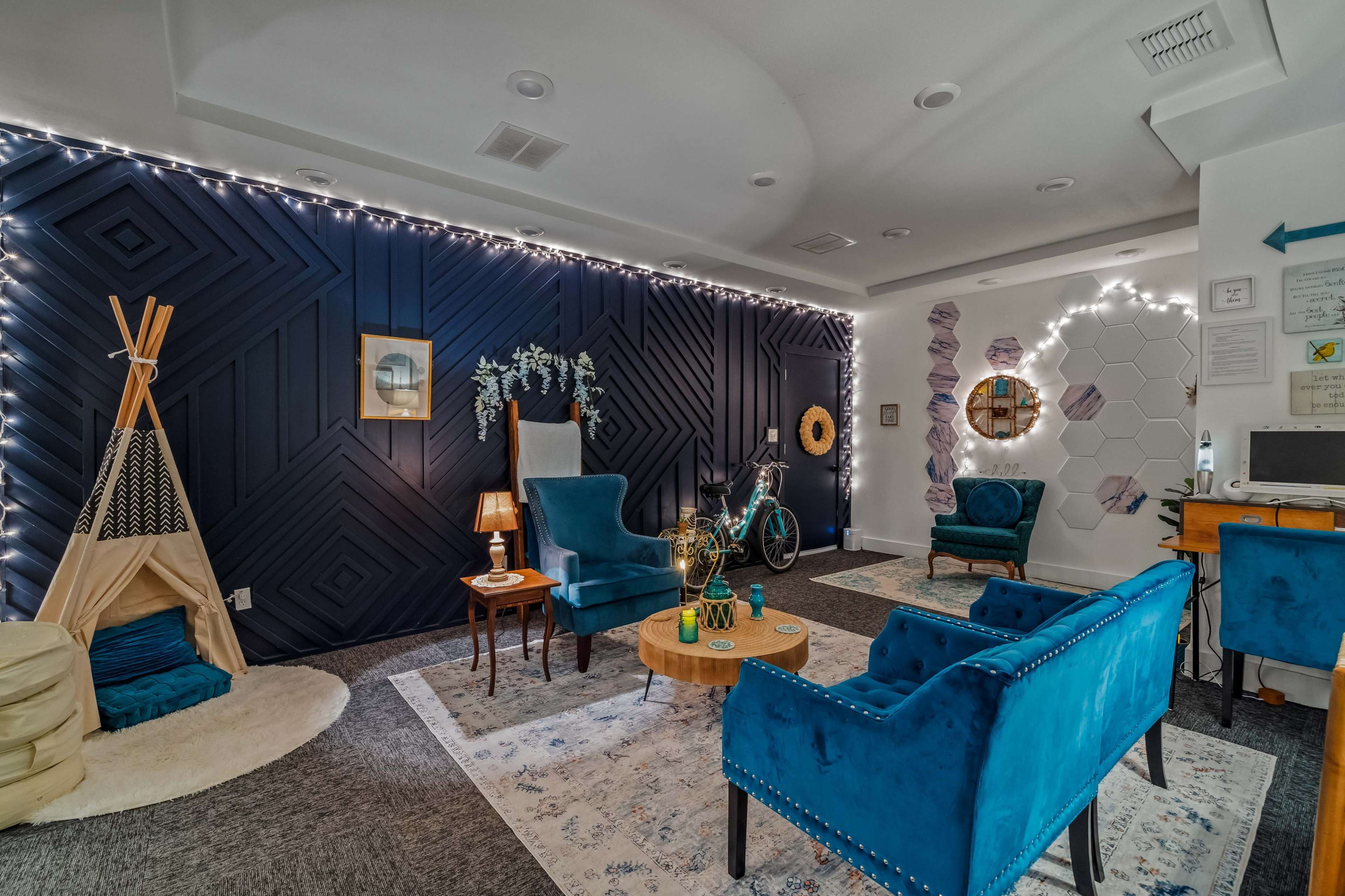 The image depicts a cozy room featuring blue upholstered chairs, a wooden coffee table, a bicycle, and decorative elements like plants and string lights against a patterned wall.