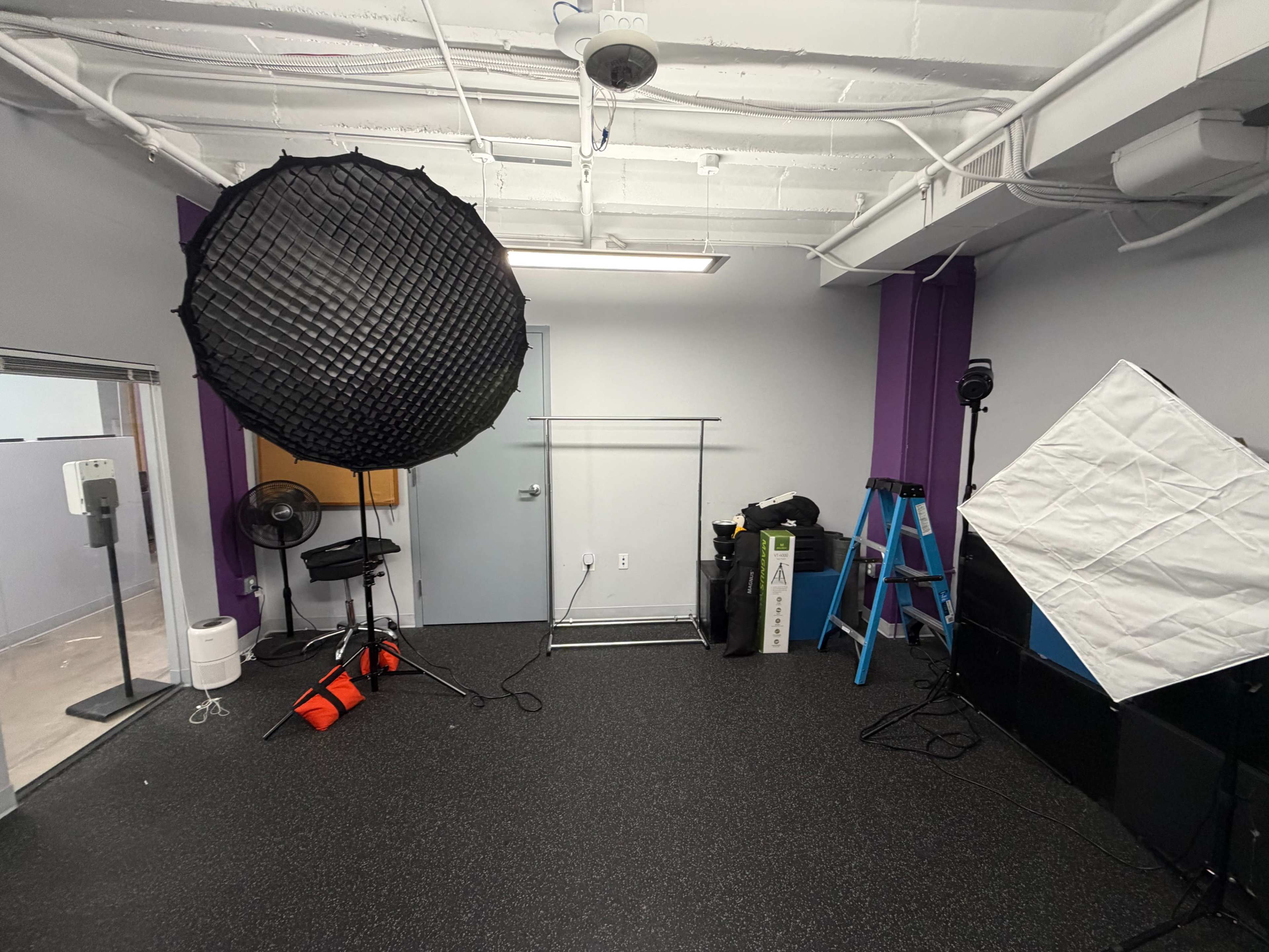 The room contains photography equipment, including a large softbox, light stands, a backdrop holder, and a ladder, all set against a plain, dark floor and colorful walls.