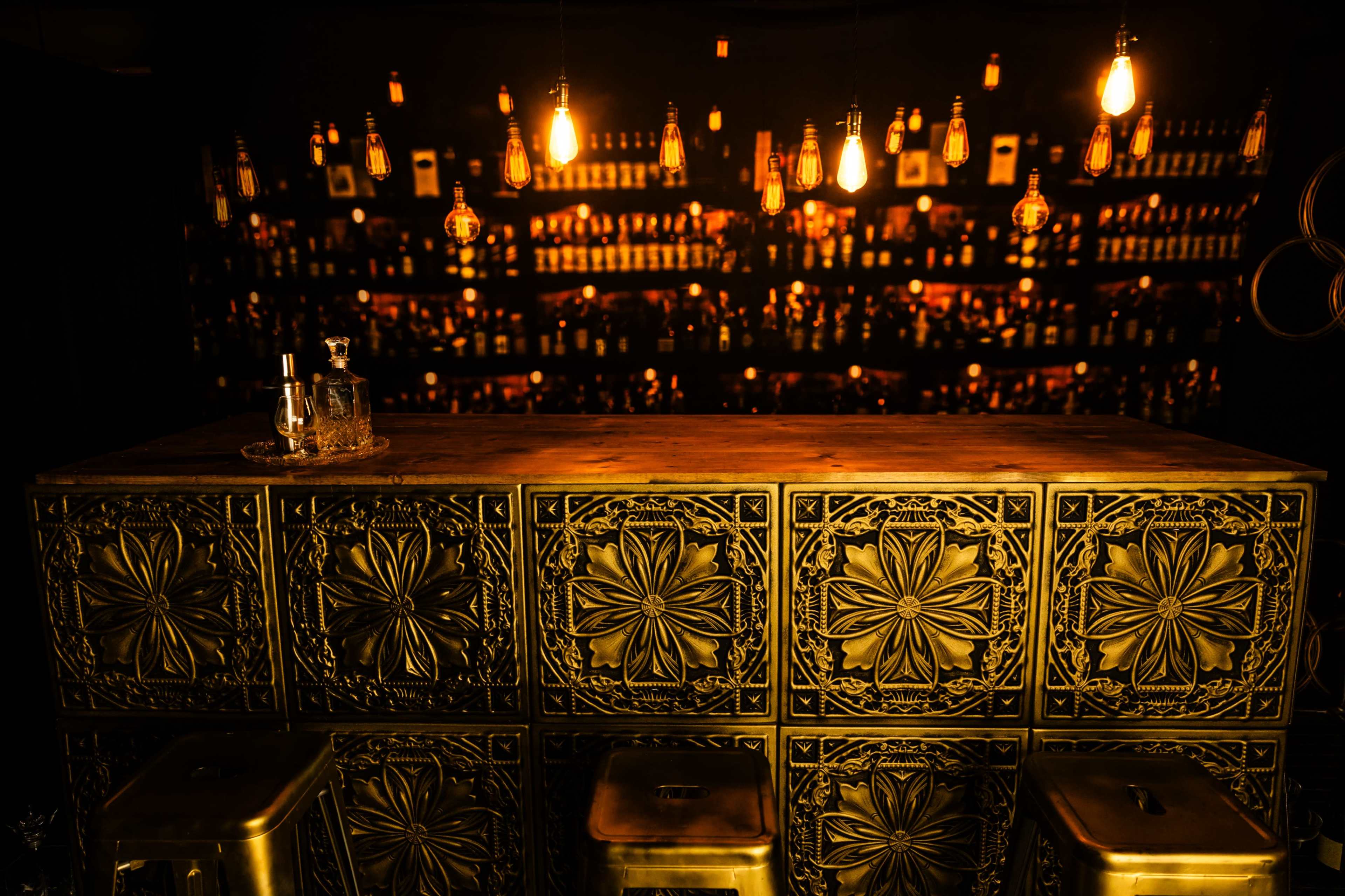 A decorative bar with an ornate metal front is illuminated by hanging pendant lights, while shelves in the background display a variety of bottles.