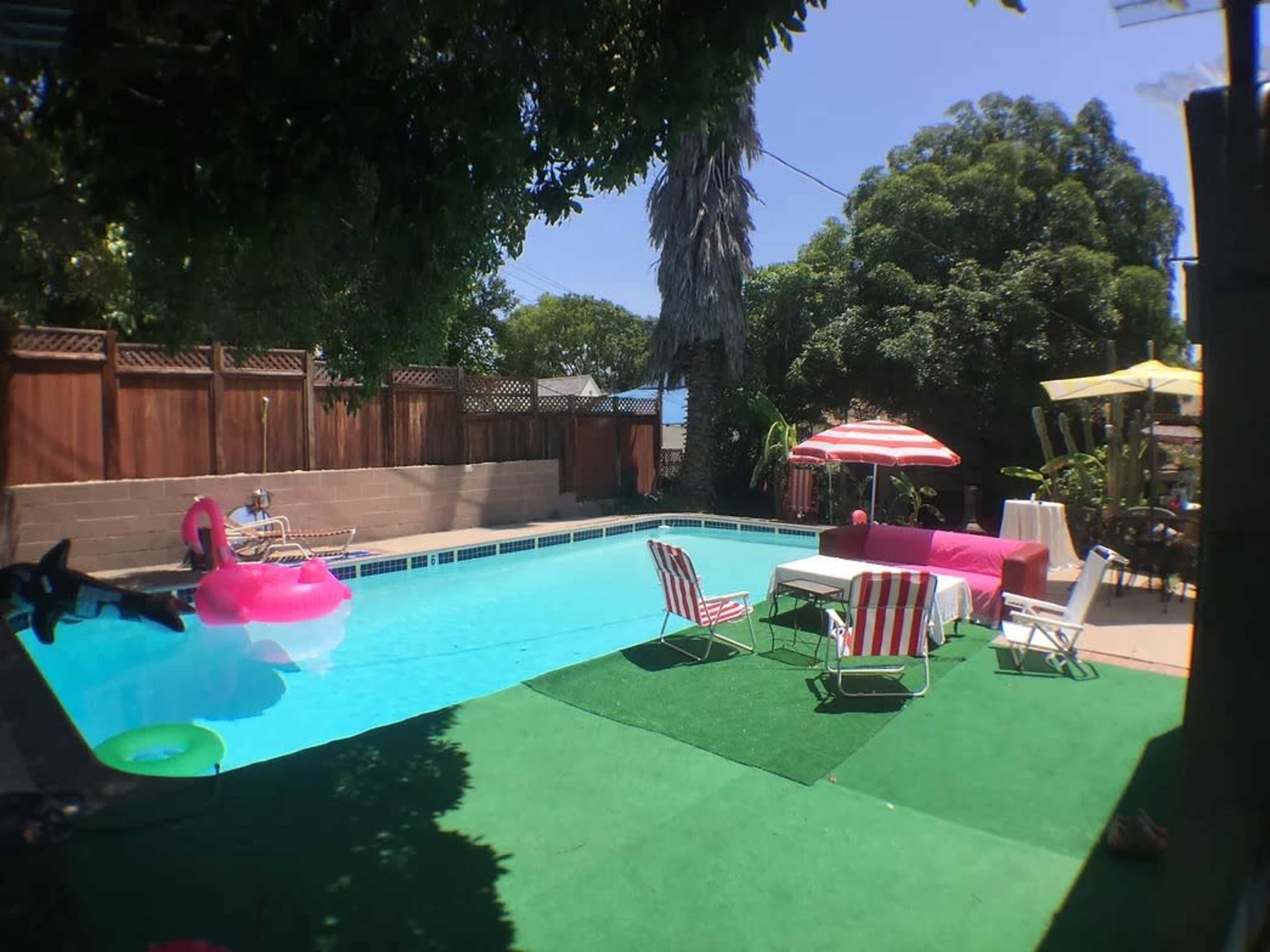 A backyard pool surrounded by lounge chairs, a pink float, and shade provided by umbrellas and trees.
