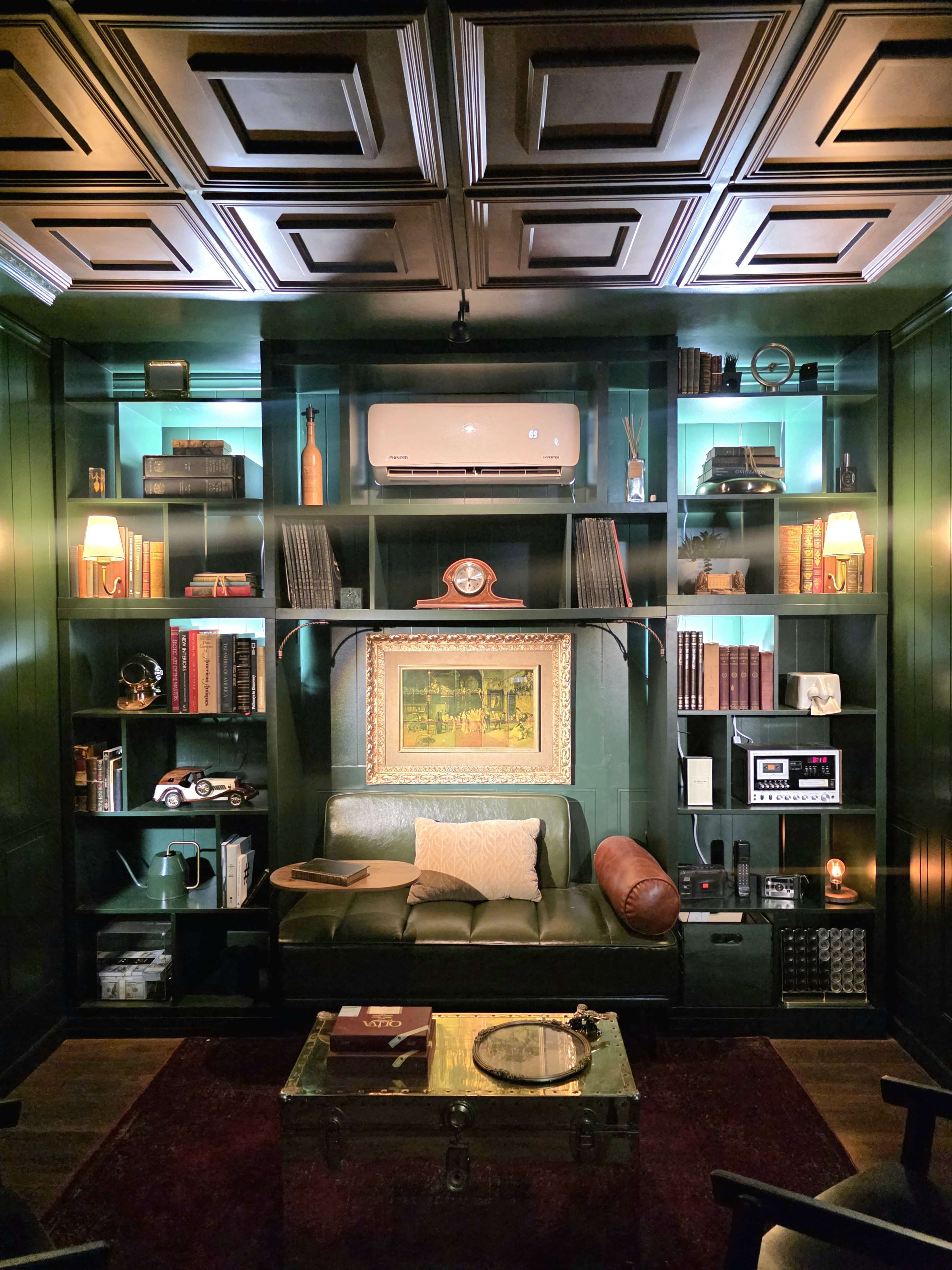 The image features a cozy library-like room with dark green walls, a bookshelf filled with books, decorative items, and a comfortable seating area.