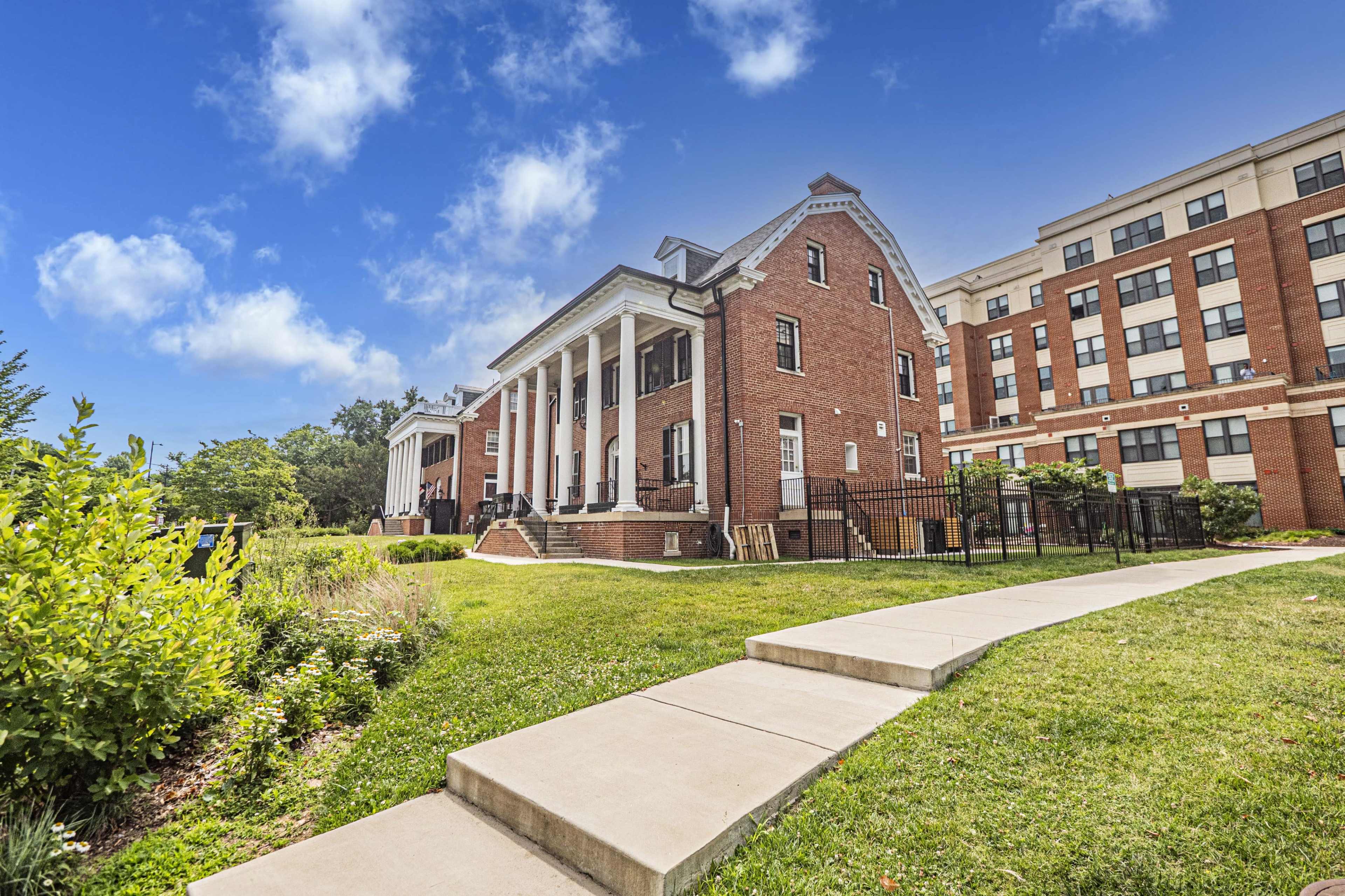 A brick building with white columns is surrounded by green grass and trees, with a concrete pathway leading up to it, under a clear blue sky.