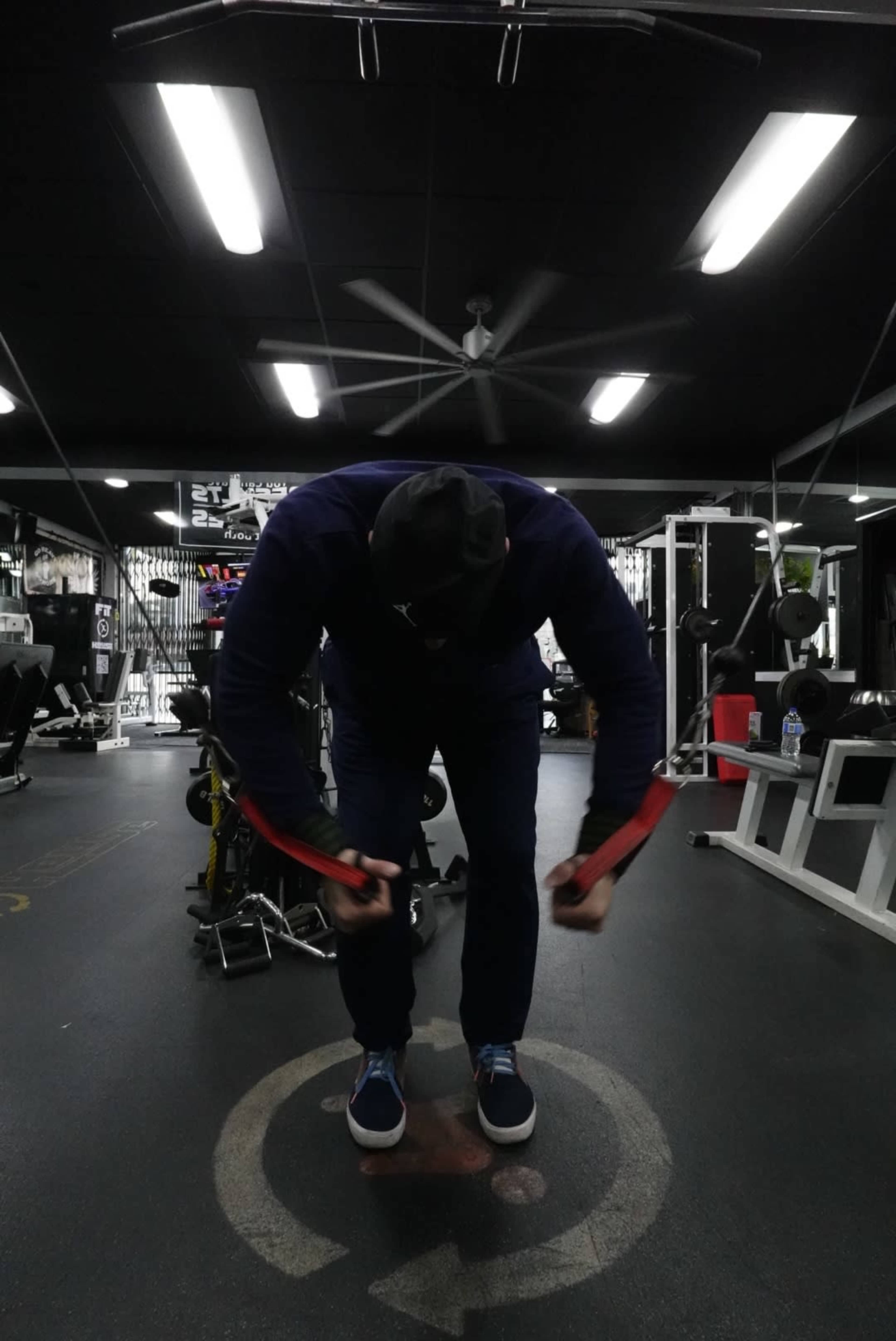 A person in a gym is performing a resistance band exercise while standing on a circular logo on the floor.