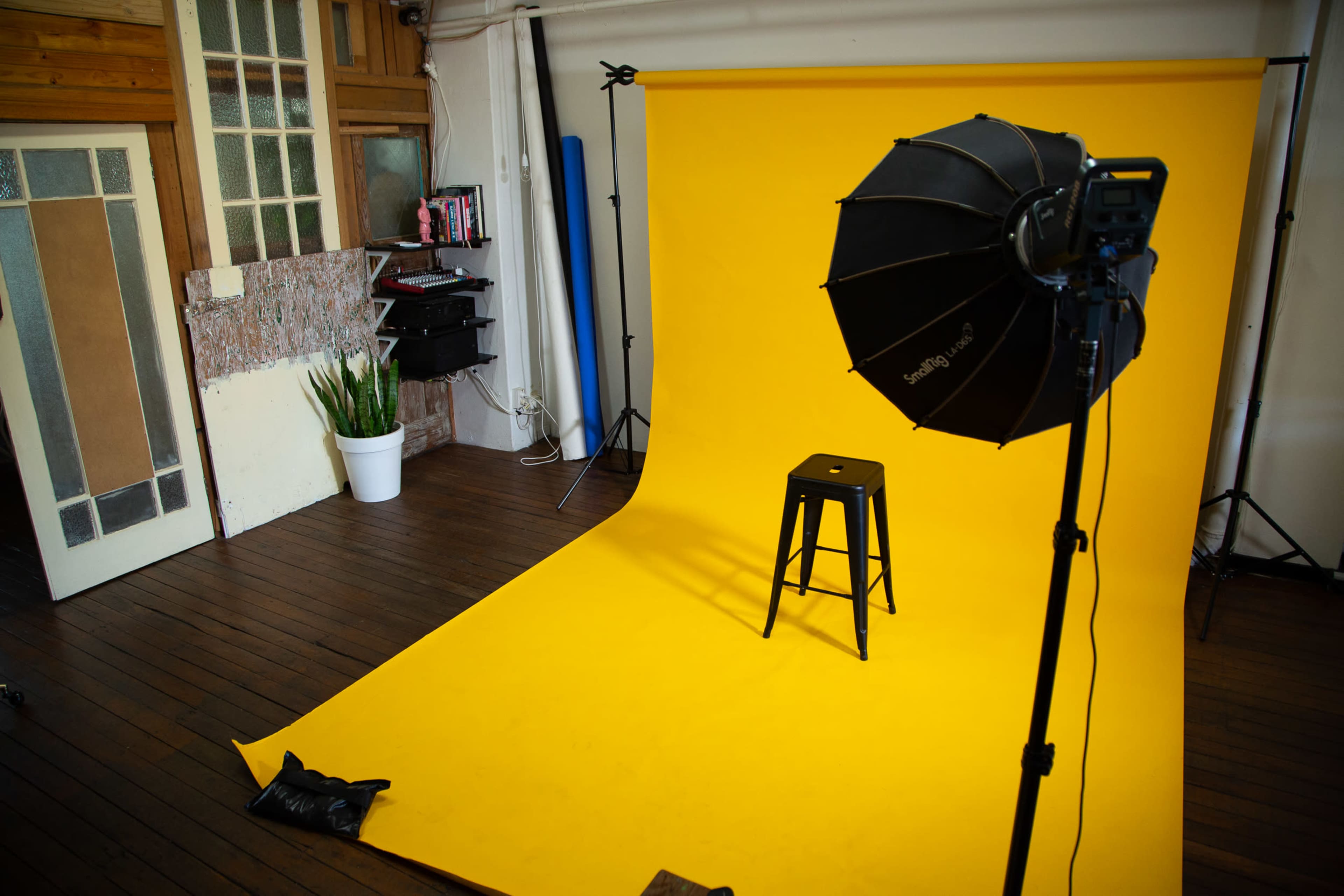 A photography studio is set up with a bright yellow backdrop, a black stool in the center, and a large softbox light positioned to the side.