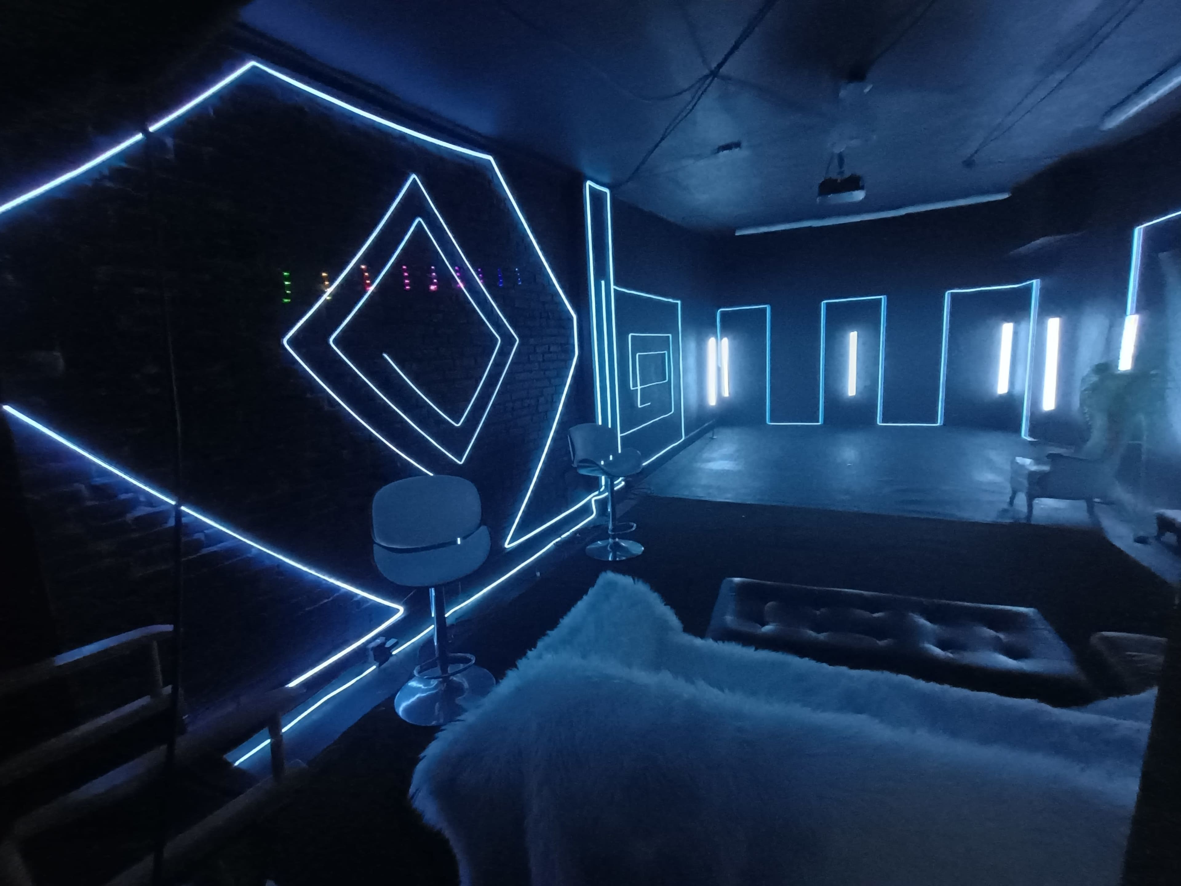 The image shows a modern interior space with glowing blue LED lights outlining geometric shapes on the black walls, along with two white bar stools and a plush seating area.