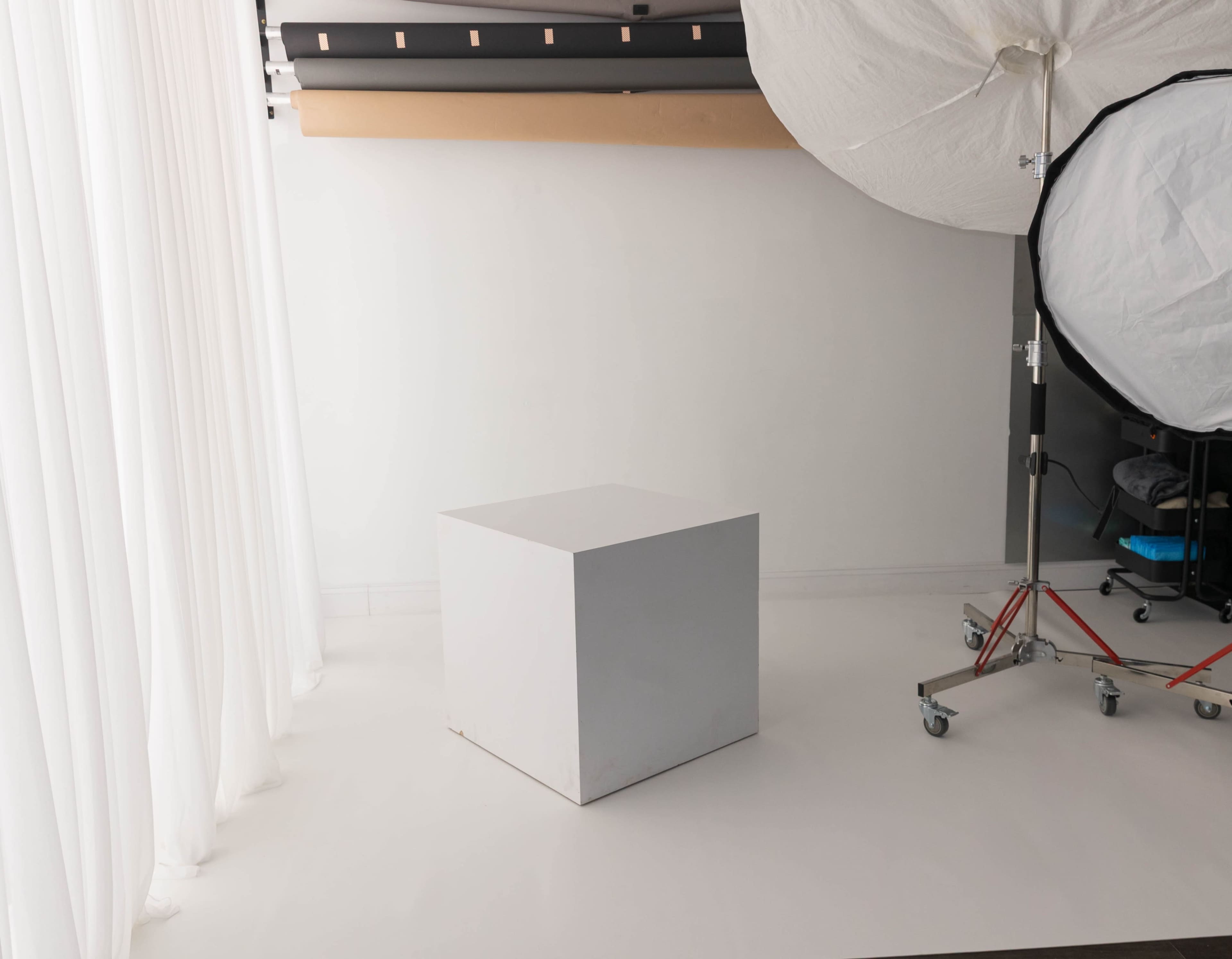 A white cubic object is placed on a light-colored floor in a studio setting, with soft drapes and a light source in the background.