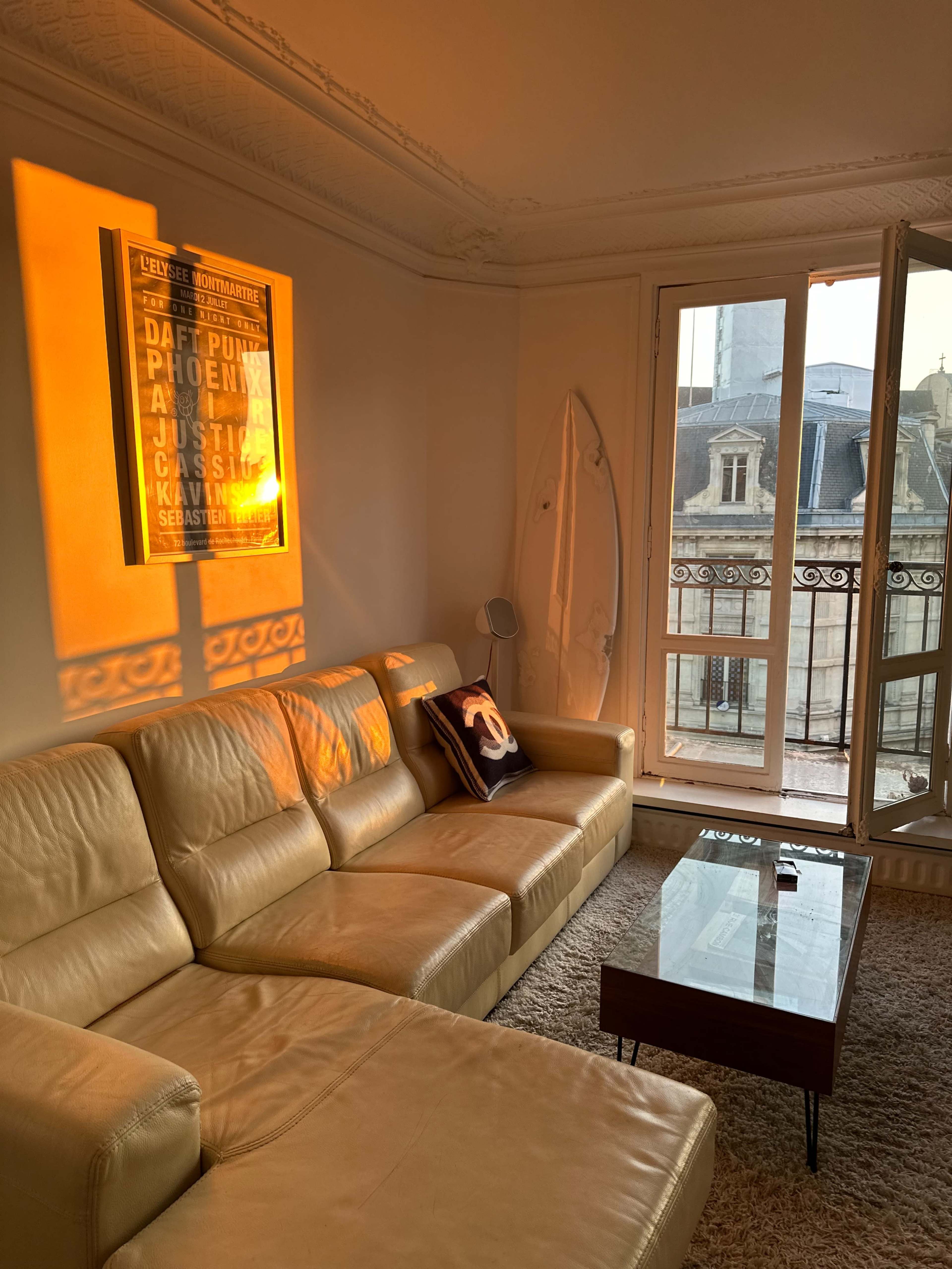 The image shows a cozy living room with a leather sectional sofa, a coffee table, and a surfboard leaning against the wall, illuminated by warm sunlight through a balcony door.