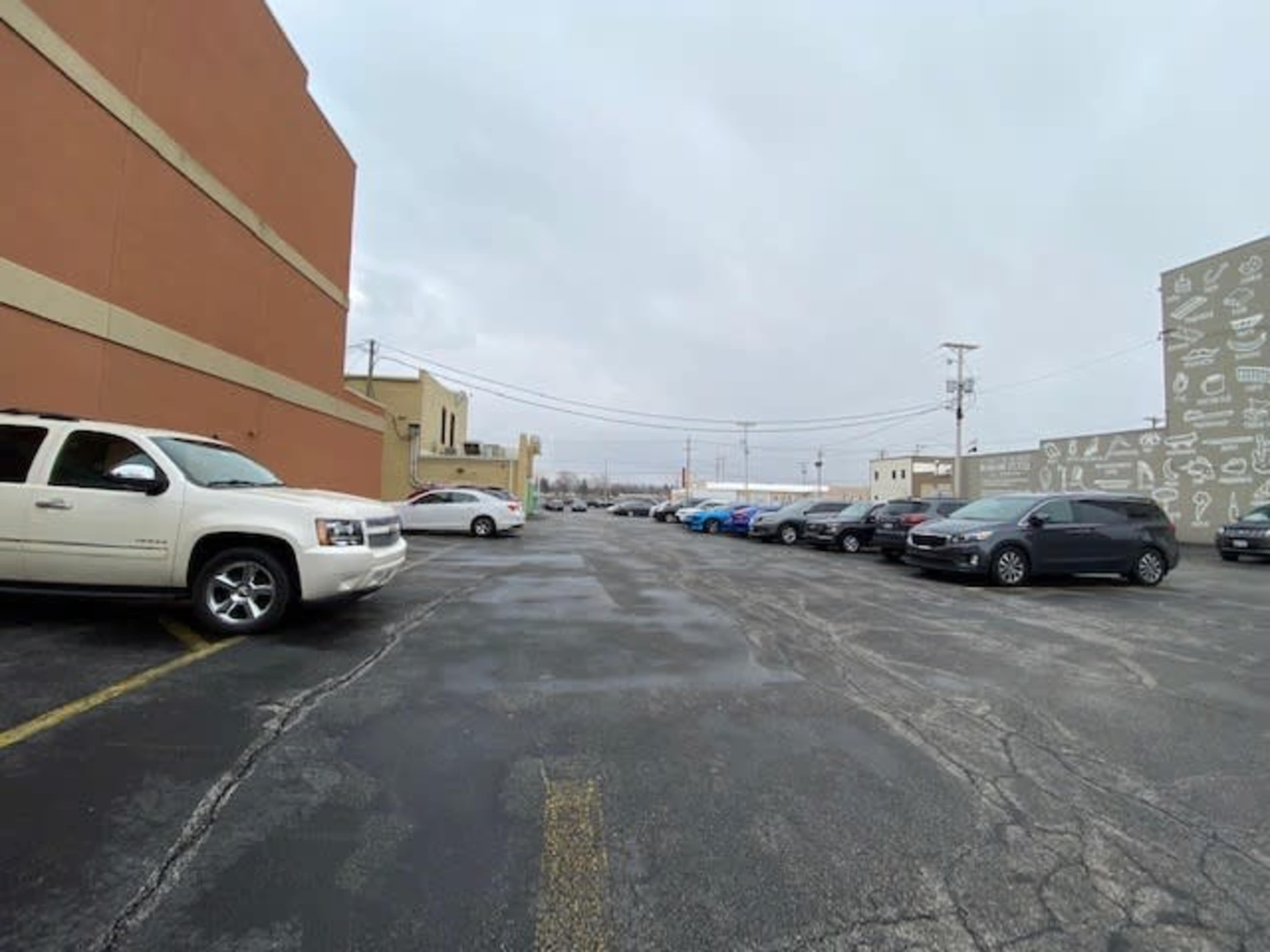 A parking lot with several vehicles lined up along the sides, flanked by two buildings under a cloudy sky.