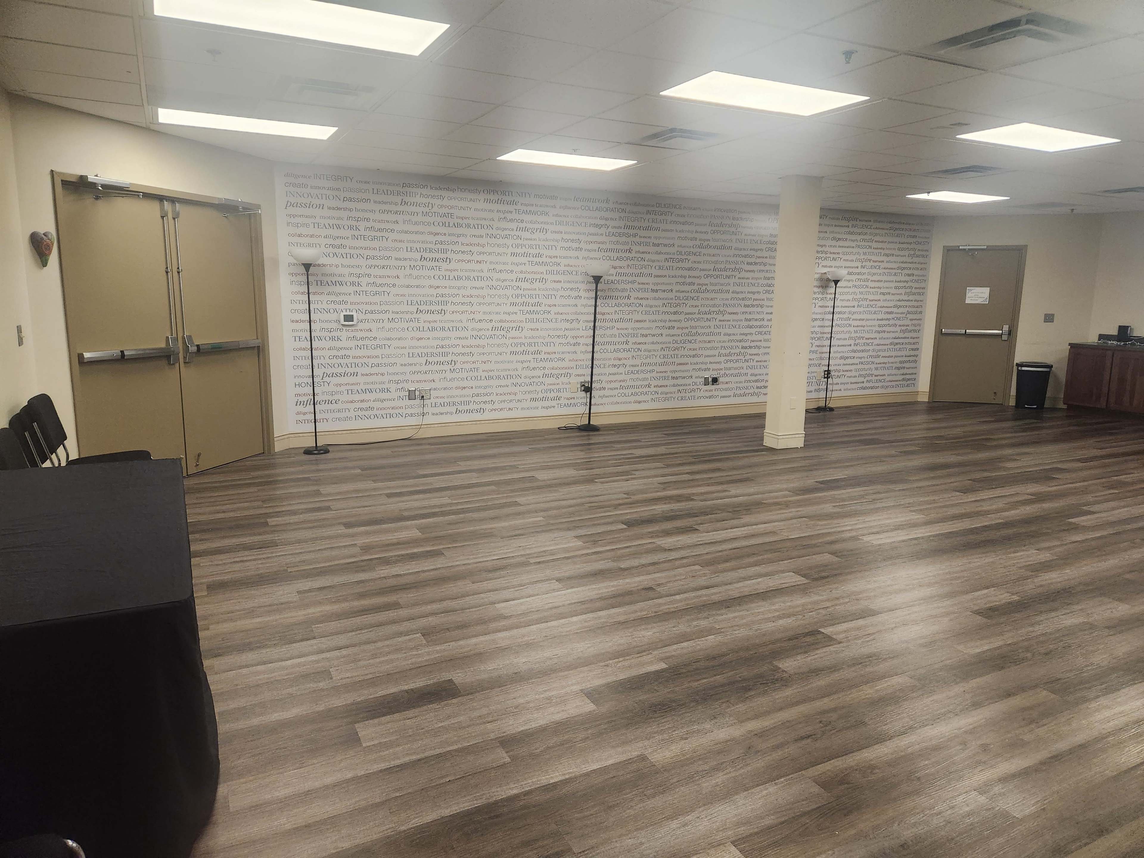 The image shows an empty room with wooden flooring, two floor lamps, and a wall covered in text, along with a table on the left side.