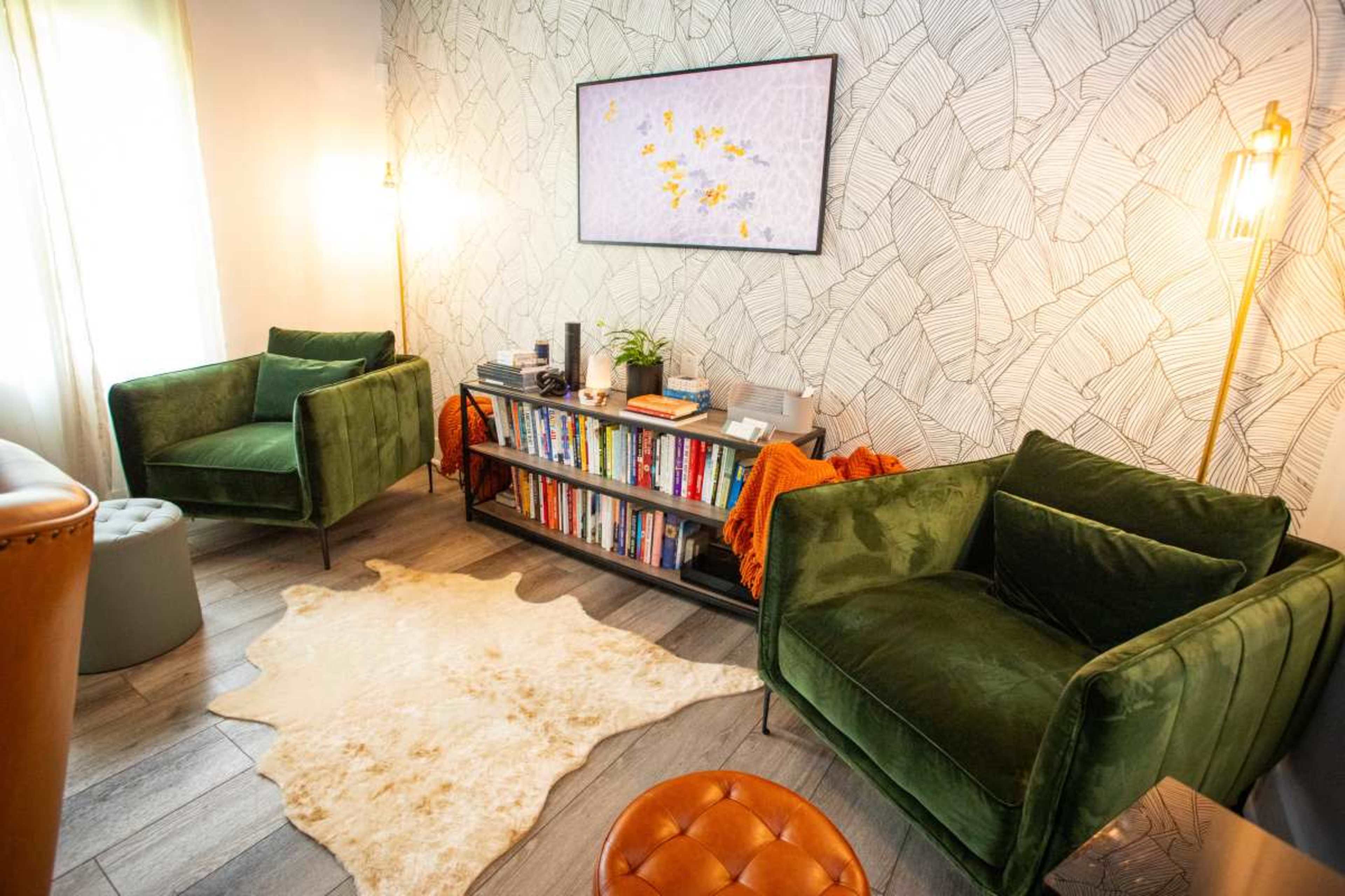 The image shows a cozy room with two green armchairs, a bookshelf filled with books, and a light-colored rug on the floor, all set against a patterned wall.