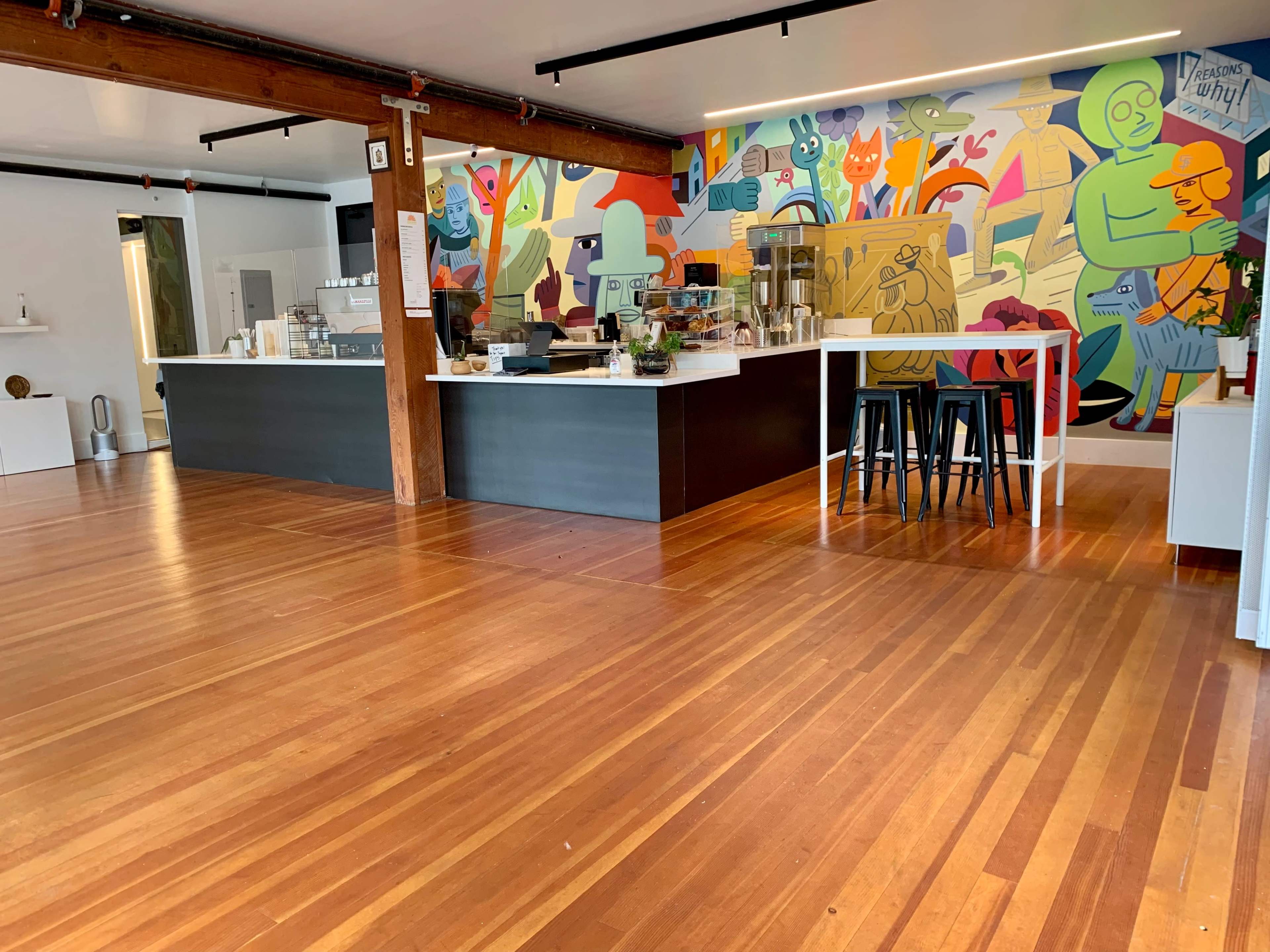 A cafe interior with a colorful mural on the wall, wooden flooring, and a service counter with seating areas.