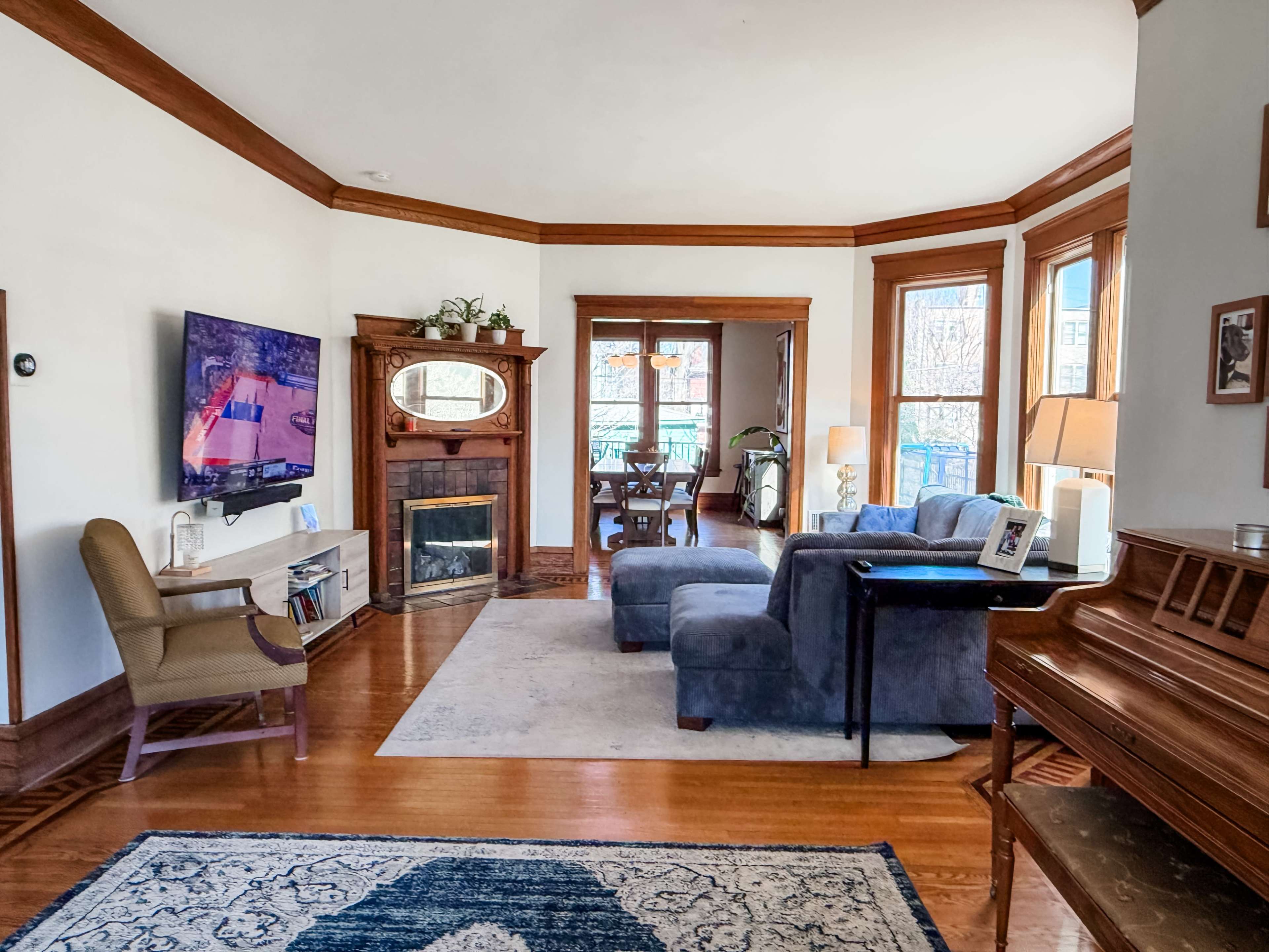 A cozy living room features a television mounted on the wall, a fireplace, a blue sofa set, and a piano, with large windows allowing natural light to enter.